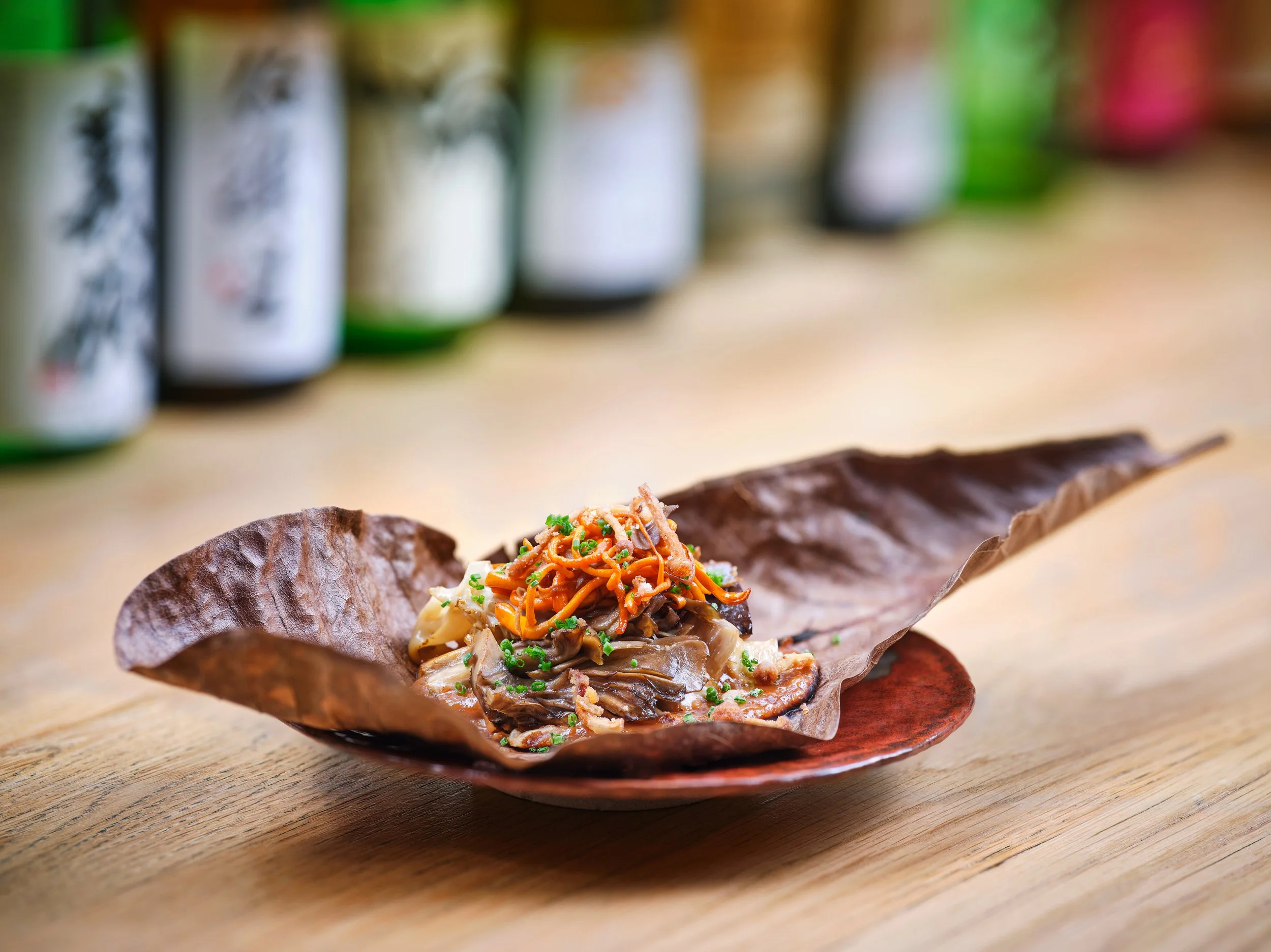 Close-up of a dish with noodles, vegetables, and chopped green herbs served in a leaf bowl on a wooden surface, with bottles in the blurred background.