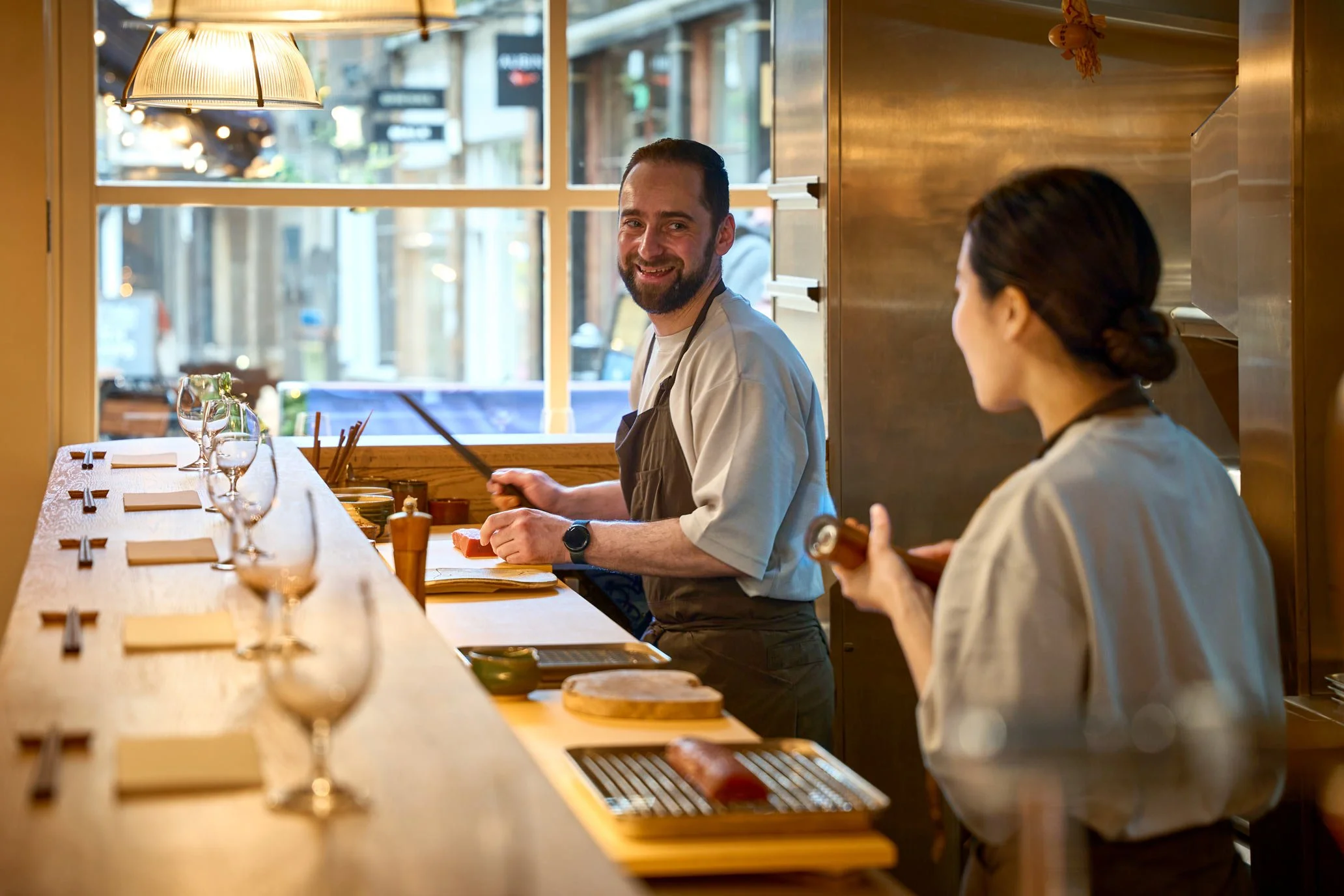 A chef with a beard smiling at a woman in a restaurant kitchen. The chef is wearing a white apron and sitting at a counter. The woman, with dark hair tied back, is holding a phone. The kitchen has wooden panels, a large window, and a hanging lamp, with a table set with glasses and utensils in the foreground.