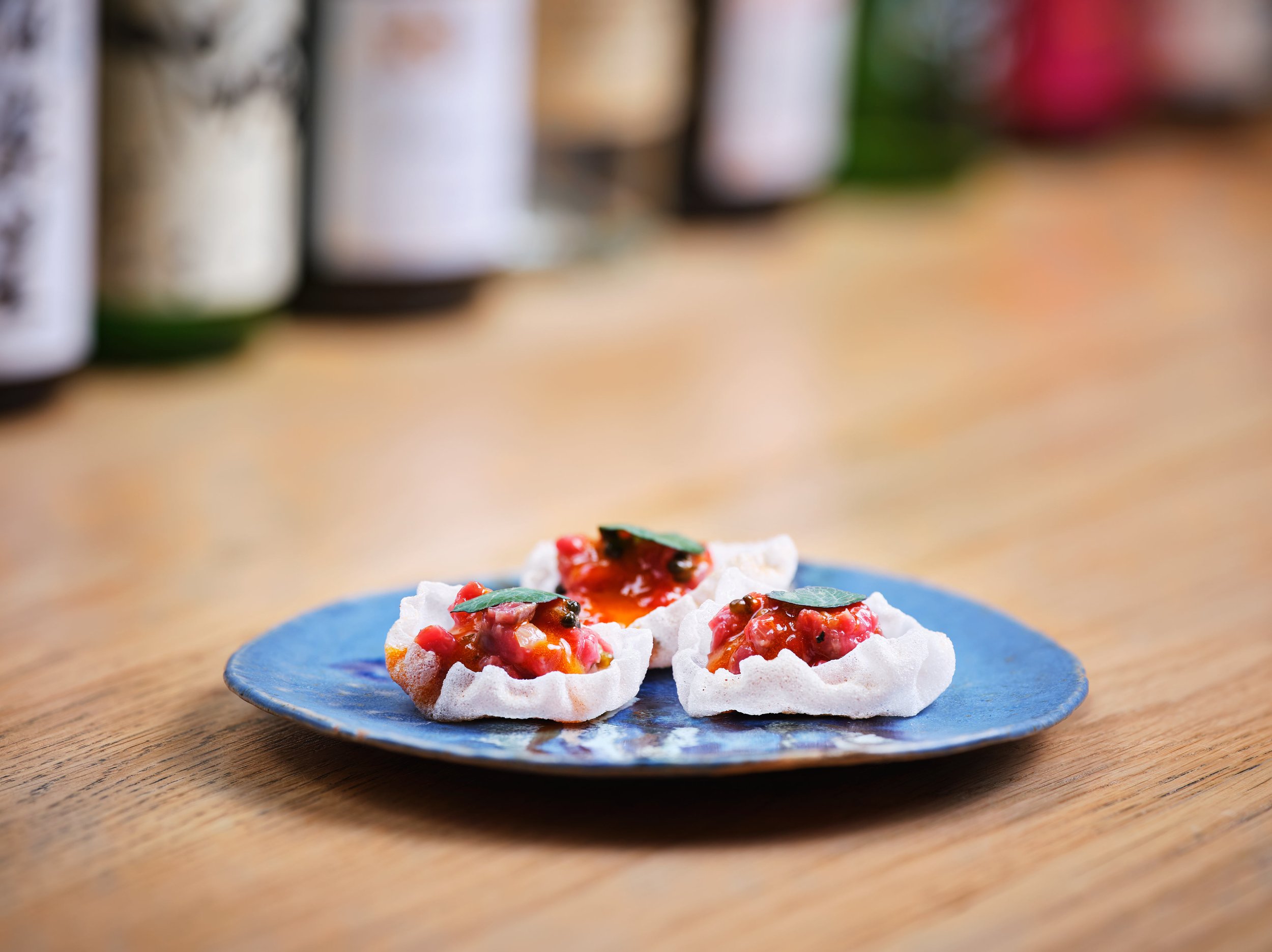 Close-up of meringue cookies topped with red fruit and green leaves on a blue plate, with blurred bottles in the background.