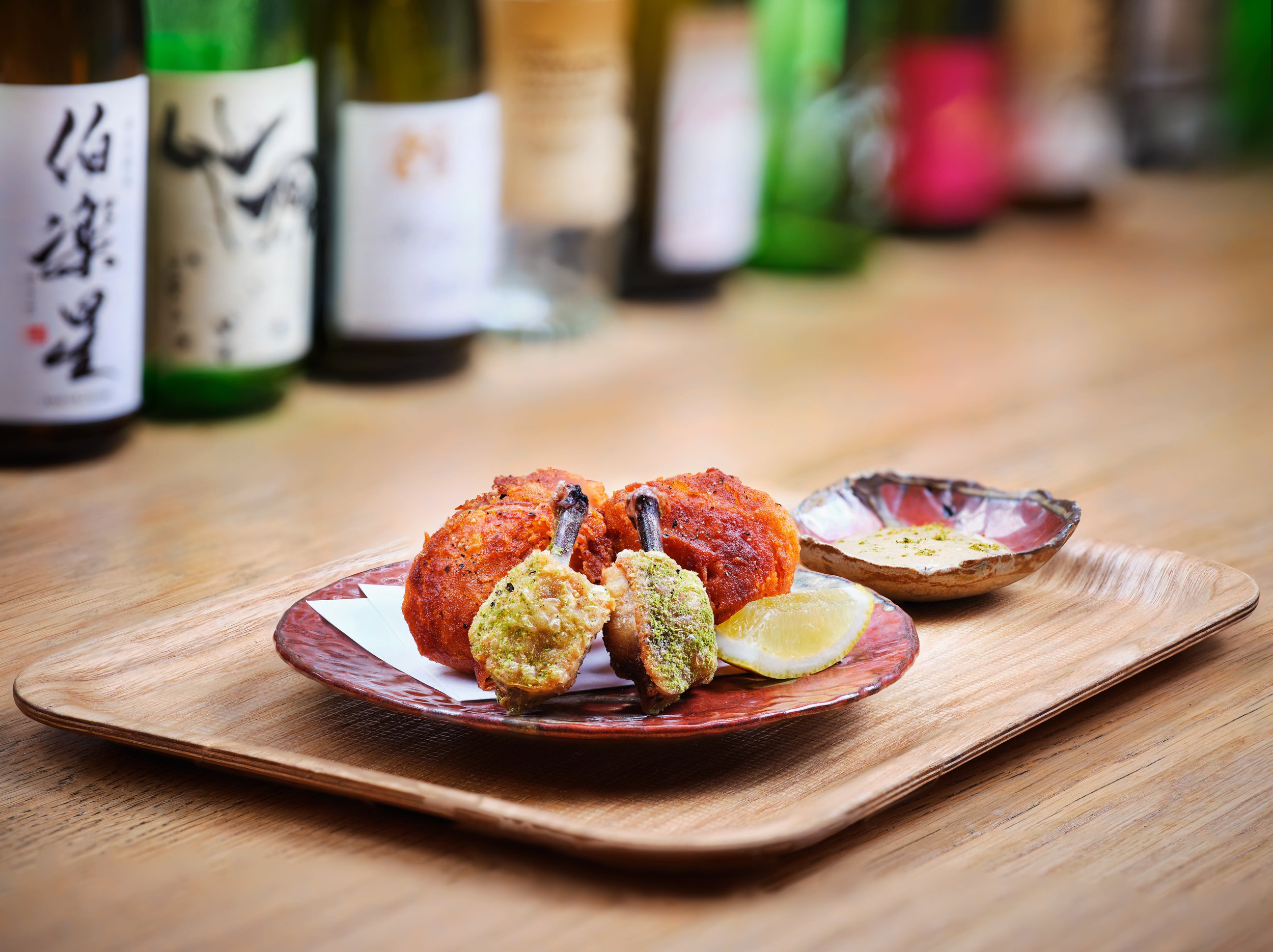 Close-up of a plate with fried skewers and lemon slices on a wooden cutting board, with bottles of sake or similar beverages blurred in the background.