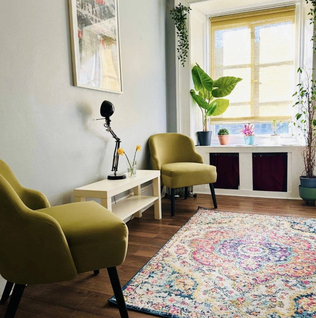 Cozy living room corner with two green armchairs, a white side table with a black adjustable desk lamp and yellow flowers, a colorful area rug, potted plants on the window sill, and framed artwork on the wall.