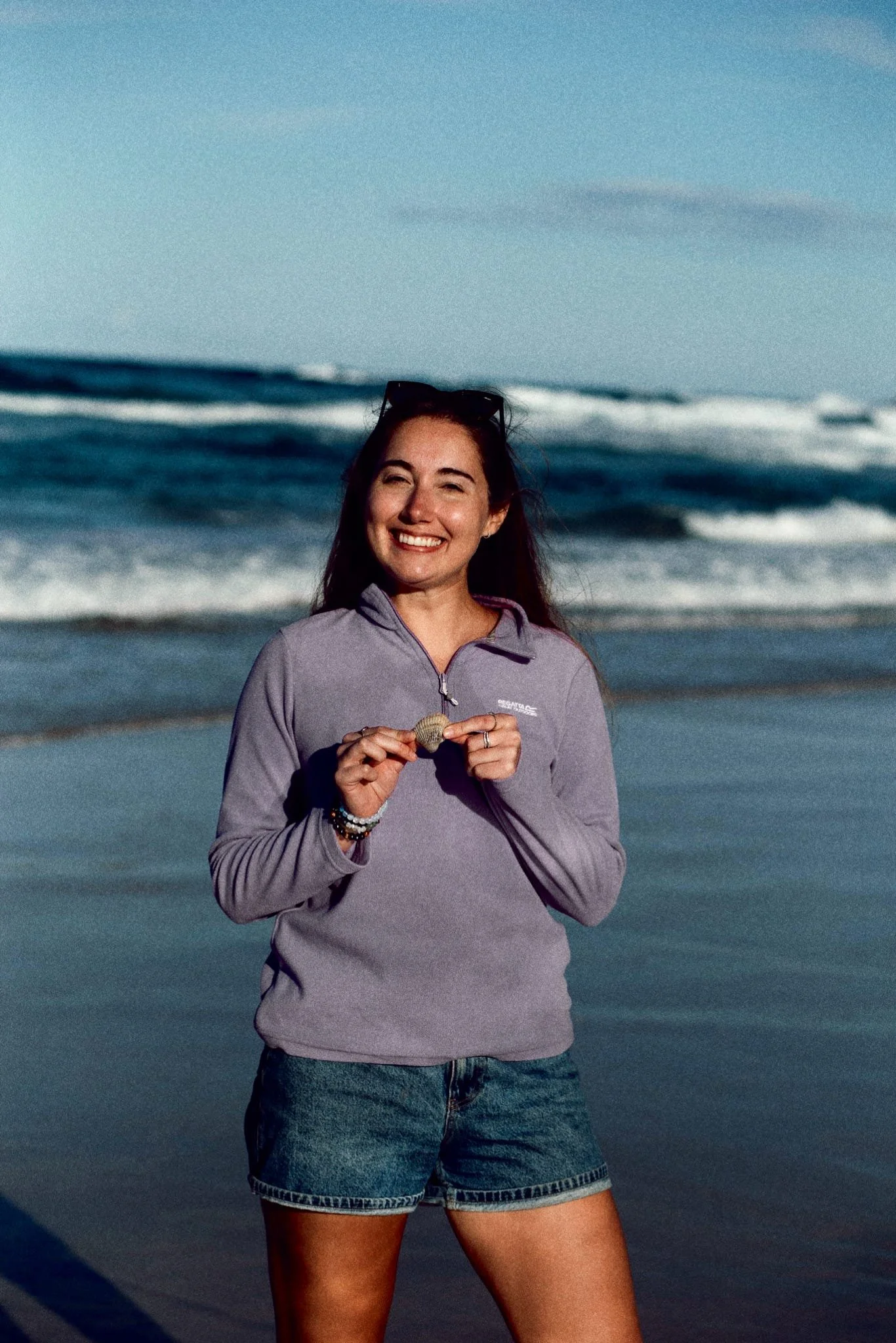 A woman smiling at the camera standing on a beach with ocean waves in the background, holding a seashell in her hands, wearing a light purple jacket and denim shorts, with sunglasses on her head.