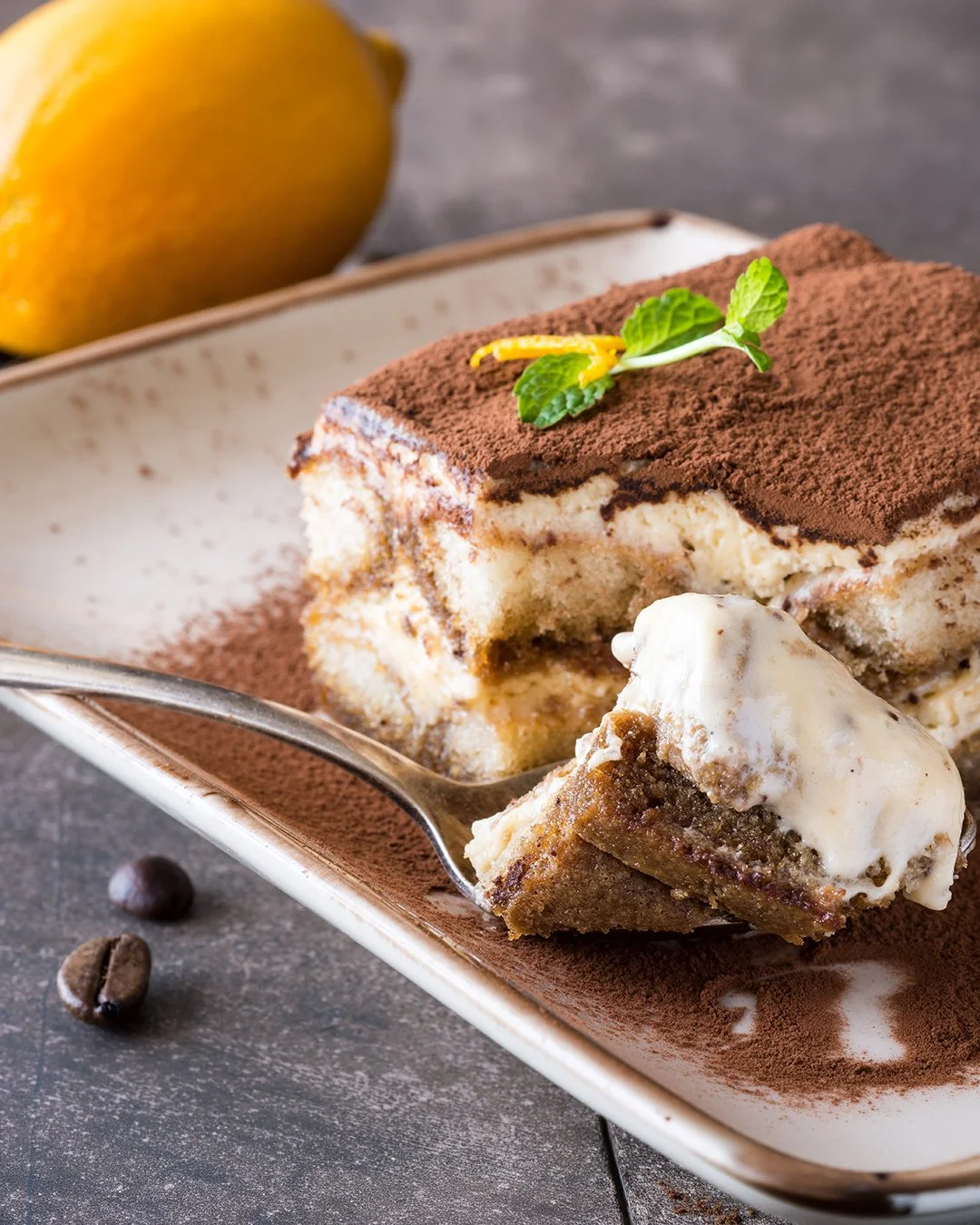 Une portion de tiramisu avec une couche de cacao en poudre, garnie de feuilles de menthe et d'un zeste de citron, servie sur une assiette longue avec des grains de café à côté.