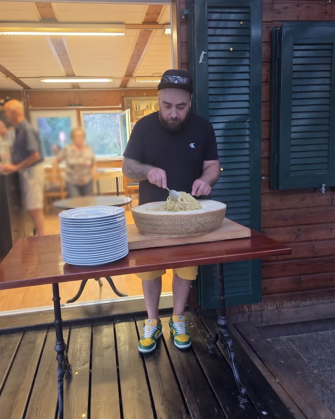 Un homme en t-shirt noir, shorts jaunes et chaussures vertes et jaunes, sert des pâtes dans un bol en bois sur une table en bois.