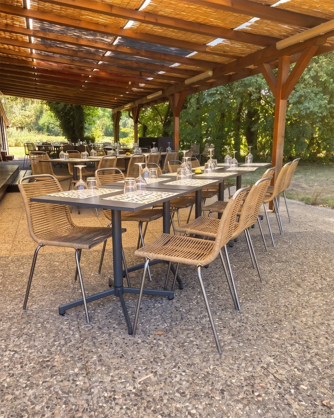 Tables de restaurant extérieures sous un auvent en bois avec des chaises en osier, prêtes pour un service, avec des verres à vin et des sets de table. Arrière-plan de verdure et de arbres.