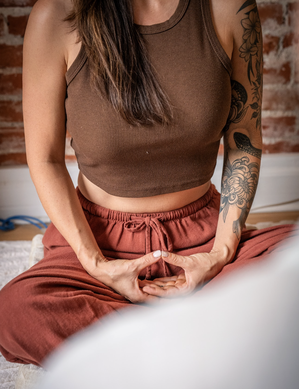 A woman practicing yoga or meditation, sitting cross-legged with hands in a mudra position, wearing a brown sleeveless top and rust-colored pants, with a detailed tattoo on her right arm.