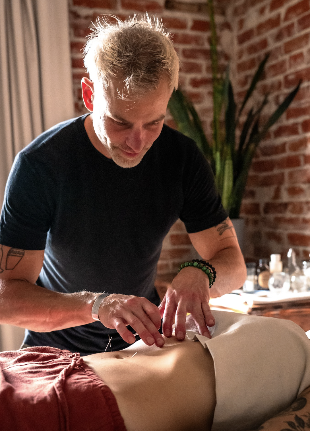 Man performing acupuncture on a person lying face down in a room with brick walls and indoor plants