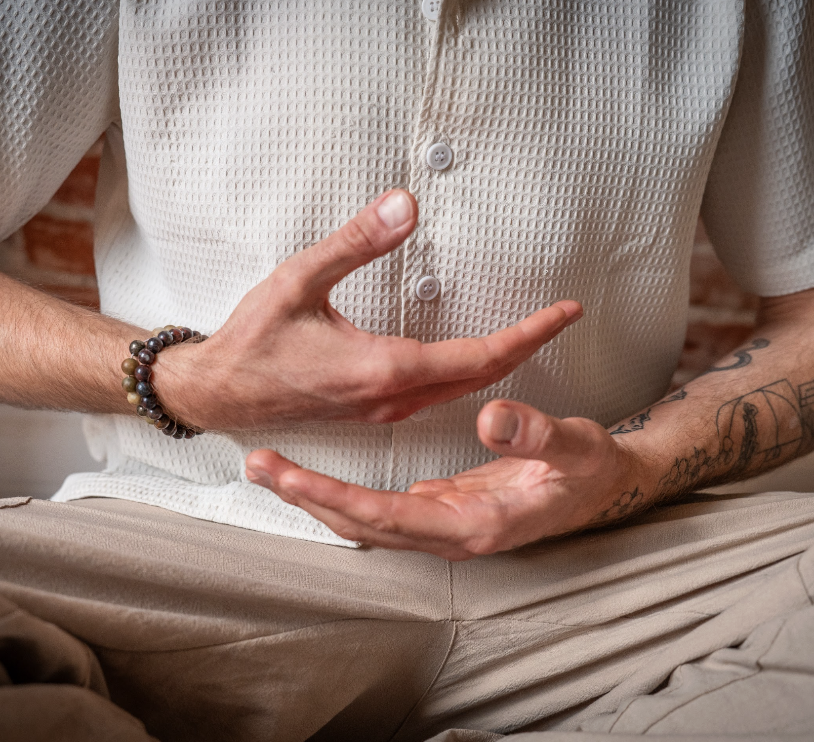 Close-up of a person crossing their arms, wearing a white textured shirt and beige pants, with tattoos on their arm and a beaded bracelet on their wrist.