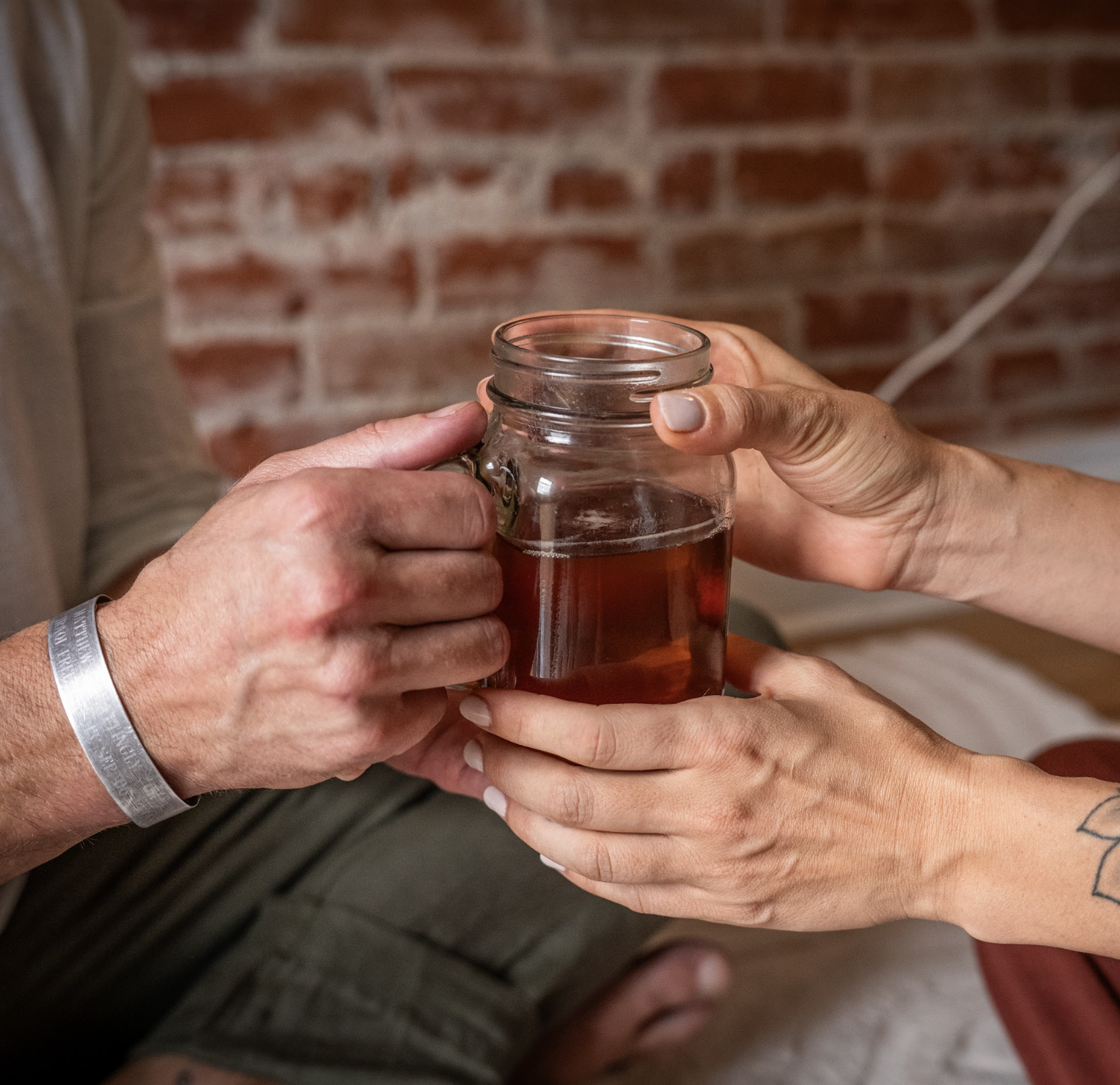 Two people clinking a mason jar with tea against a brick wall background.