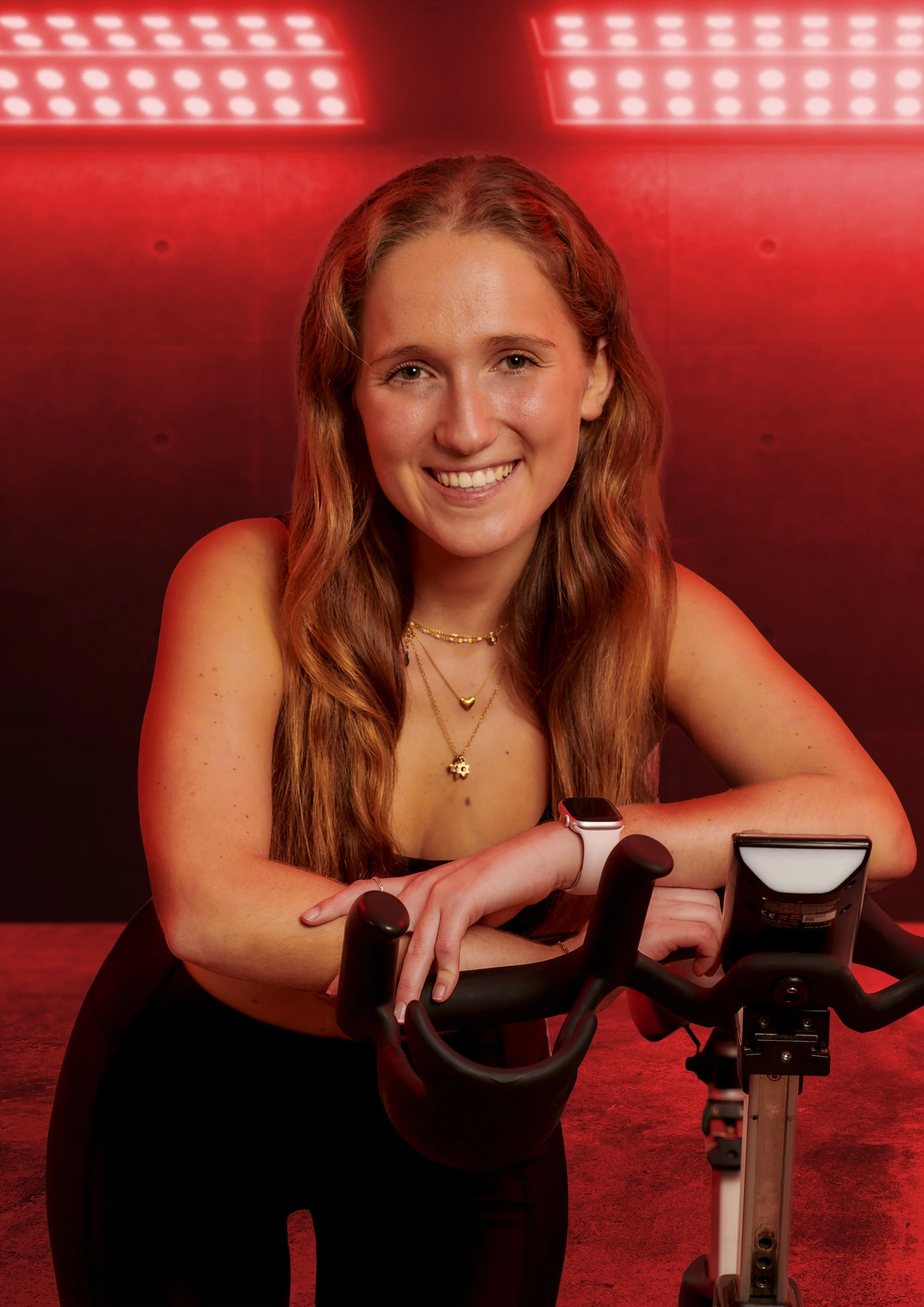 Young woman with long wavy hair smiling and sitting at a table, wearing a black top, jewelry, and a smartwatch, with exercise equipment and a red glowing background.