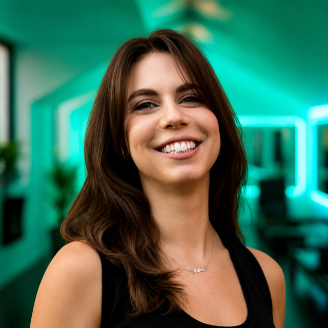 A smiling woman with long brown hair and a black sleeveless top, in a modern, well-lit room with teal and blue lighting.