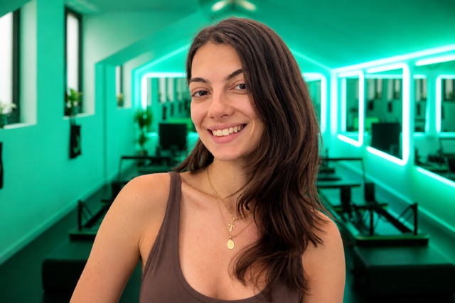 A young woman with long brown hair smiling in front of a mirrored wall with green neon lights in a modern room.