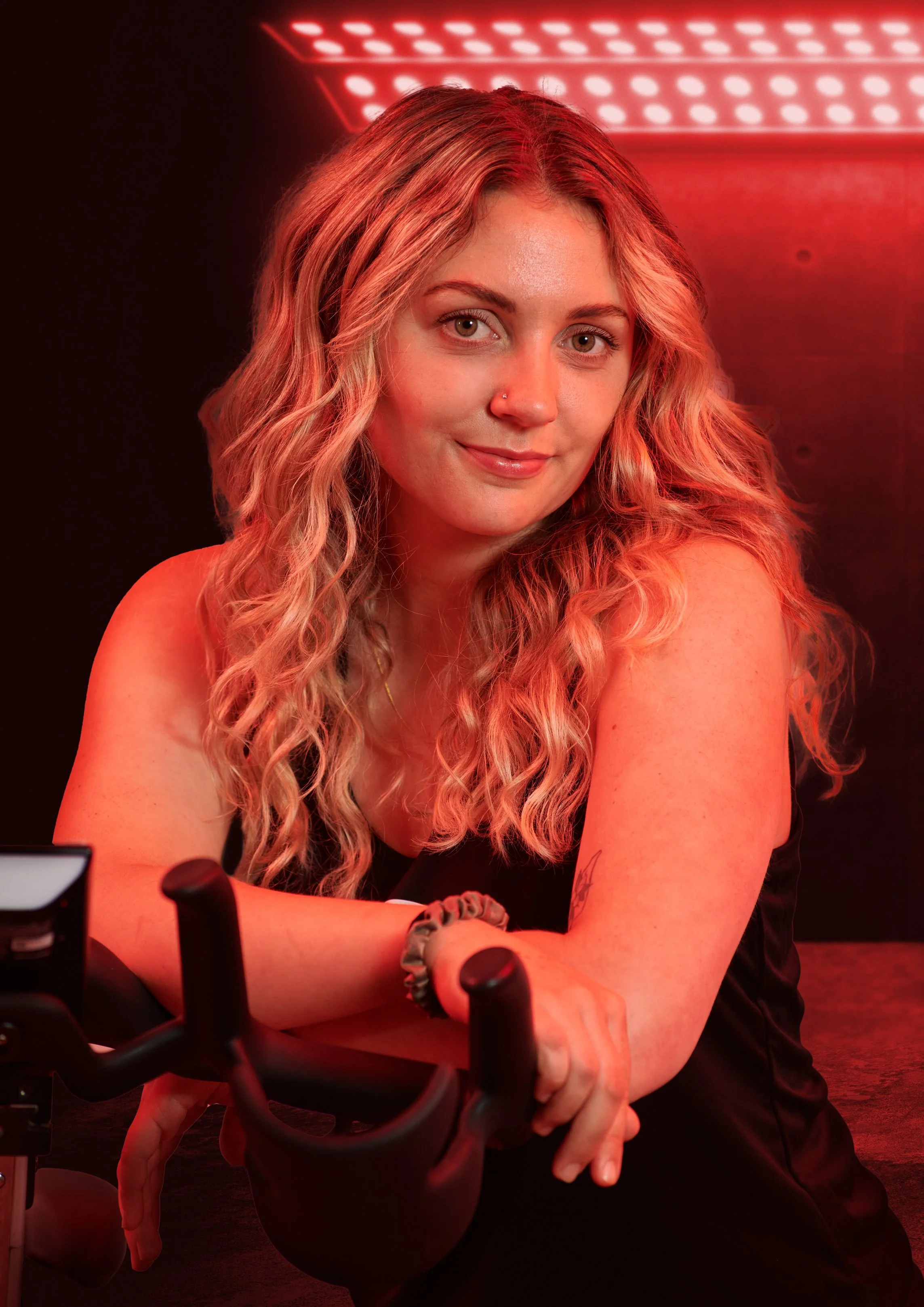 A young woman with long, curly blonde hair and a nose piercing is sitting at a table with a dark background and red lighting. She is smiling gently and looking at the camera, with her arms resting on the table. There are some electronic equipment or devices in front of her.