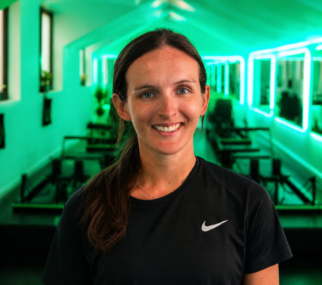 Smiling woman wearing a black Nike shirt in a room illuminated by green neon lights, with mirrors and benches in the background.