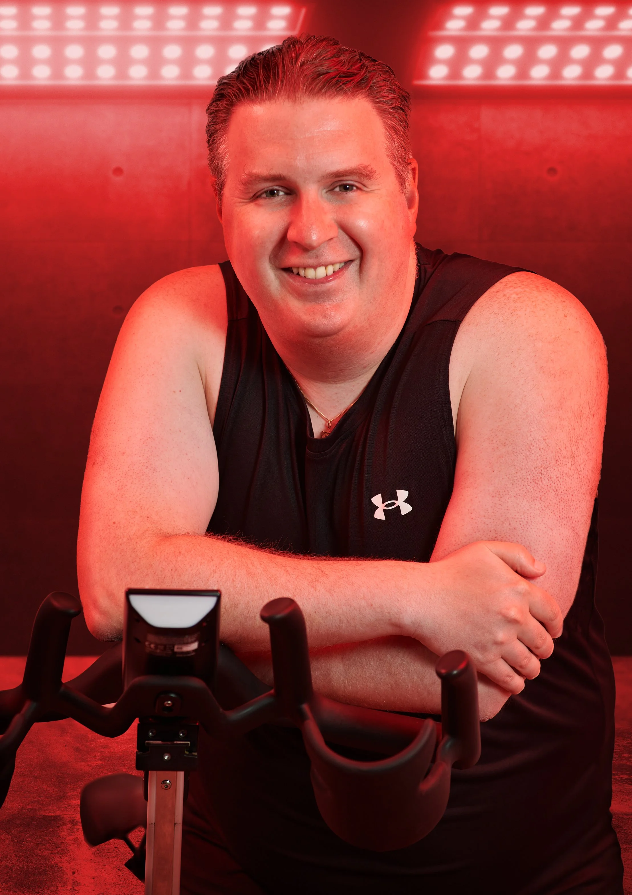 Man smiling while sitting with arms crossed, wearing a black Under Armour tank top, with a foldable exercise bike in front of him and a red LED-lit background.