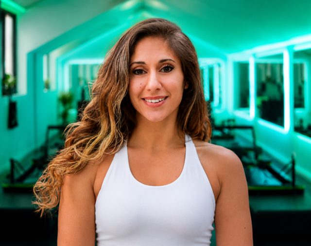 Young woman with long curly hair smiling in a brightly lit room with turquoise neon lights and mirrors.