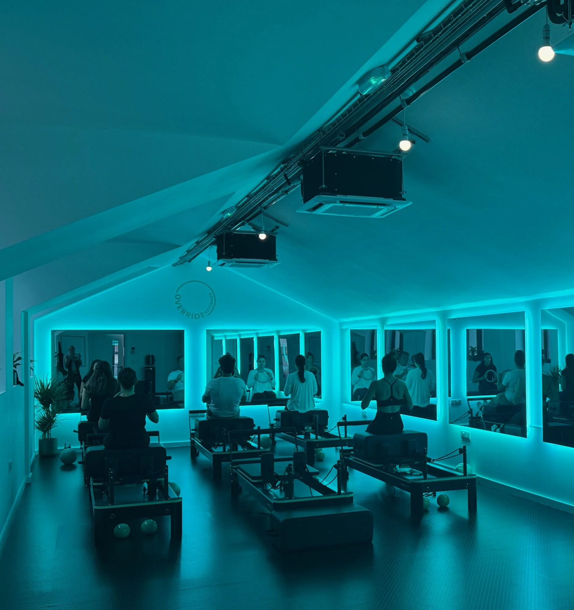 A group of people playing billiards or pool in a room with blue lighting, a mirrored wall, and ceiling lights. Pool tables are visible with cues and balls on the tables.