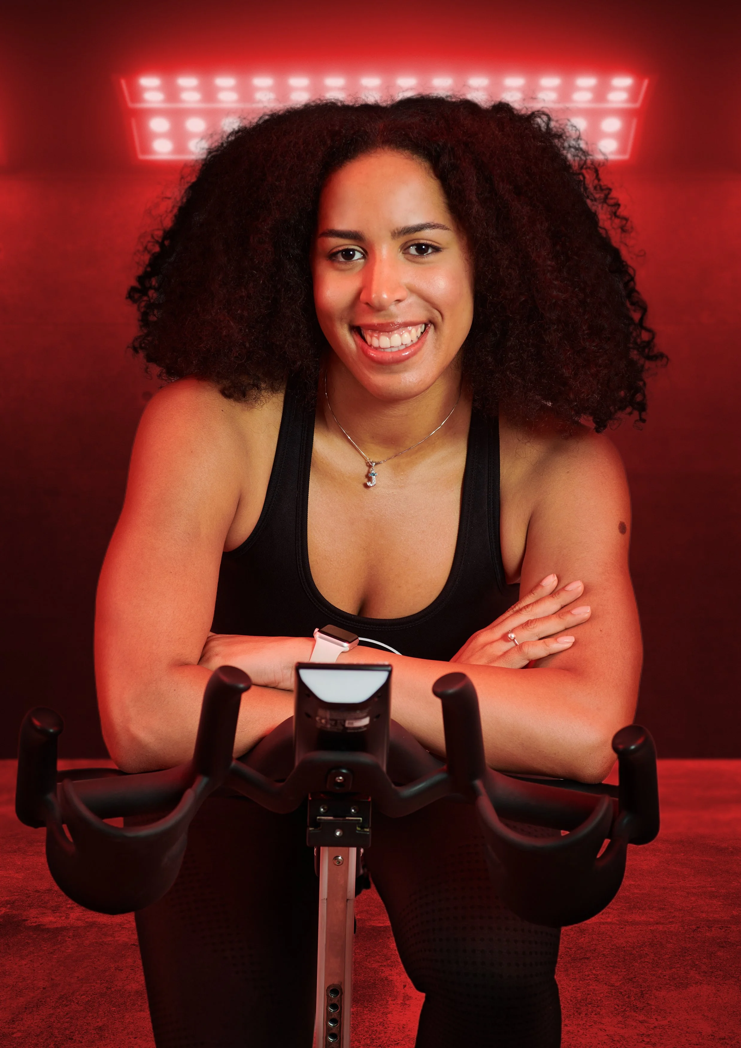A woman with curly hair smiling, sitting on a fitness bike in a red-lit room.