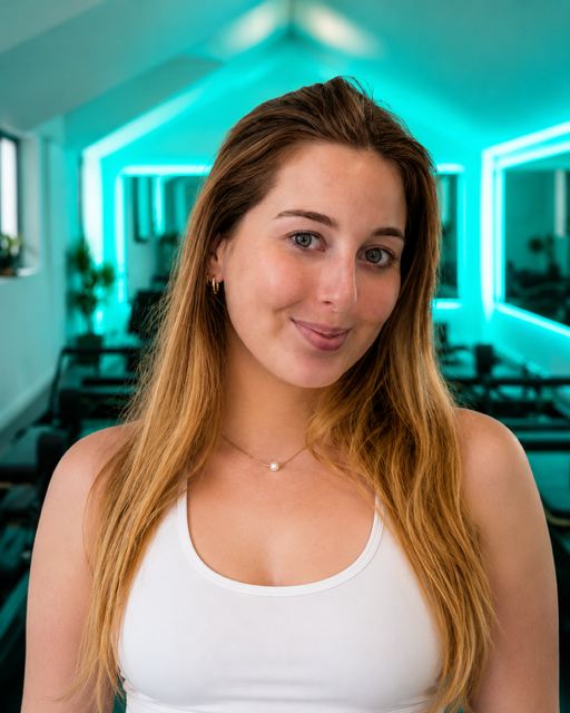A young woman with long, light brown hair and blue eyes, smiling softly, inside a room with blue neon lighting and lounge chairs.