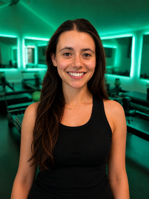 A young woman with long brown hair smiling in a gym with green lighting and exercise equipment in the background.