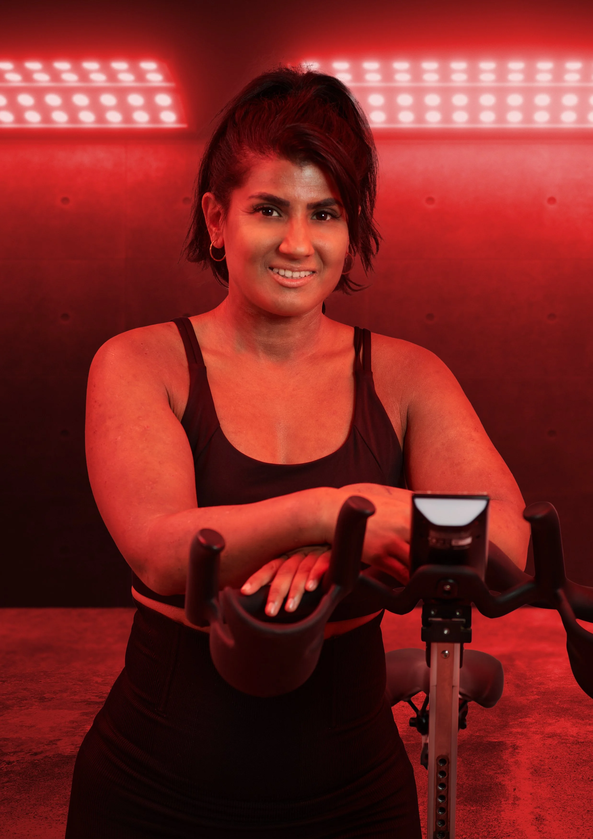 A woman with short dark hair exercising on a stationary bike in a gym with red lighting.