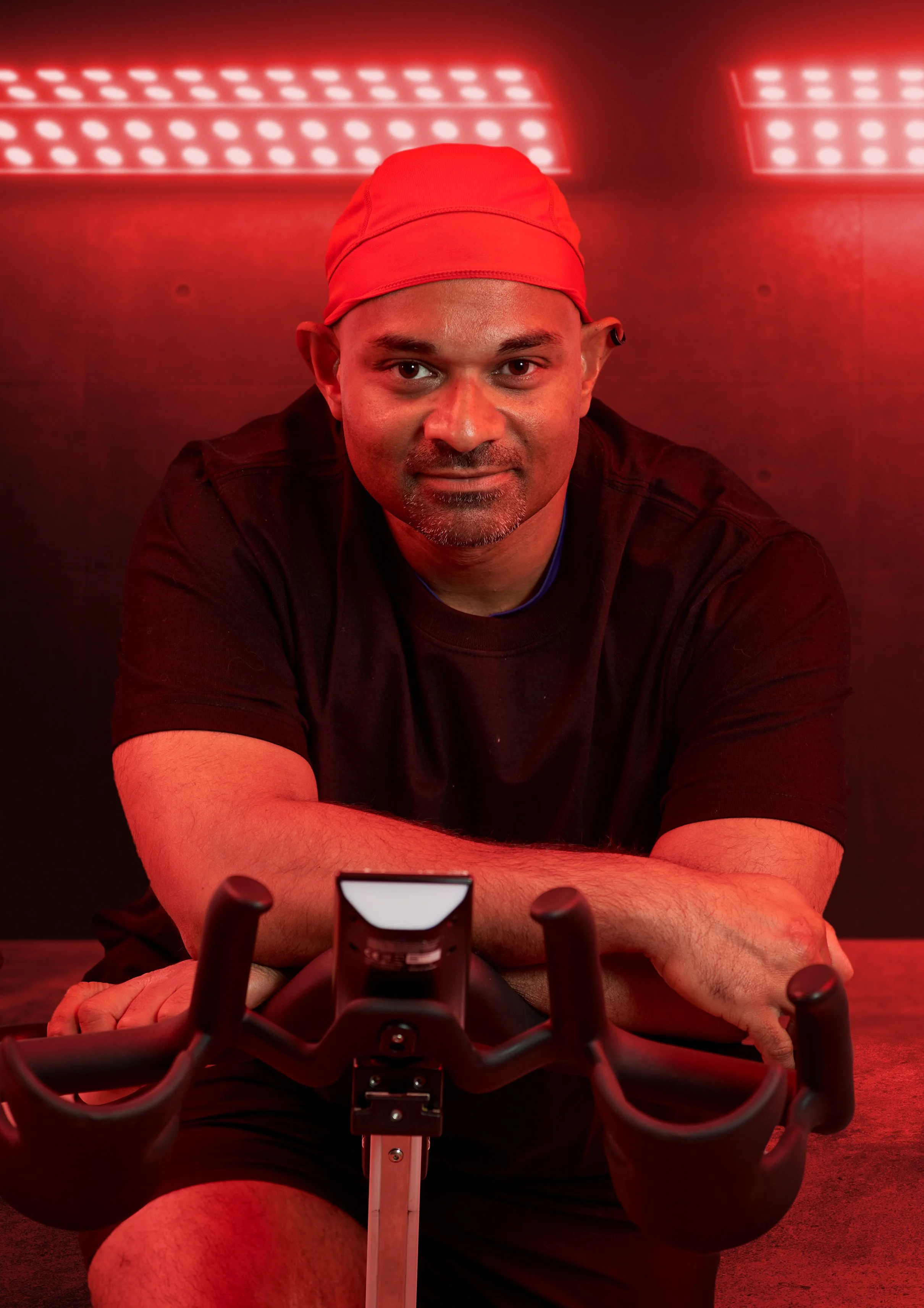 A man wearing a red cap and black shirt riding a bicycle indoors, with red lighting and a red background.