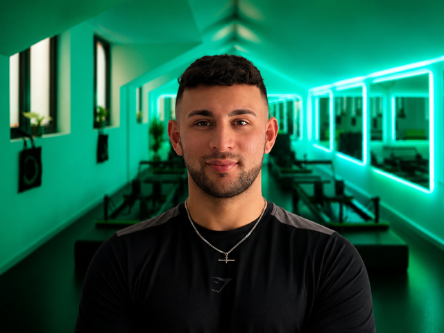 A man with short dark hair and a beard looking at the camera inside a brightly lit room with green neon lights, indoor plants, and windows on the side wall.