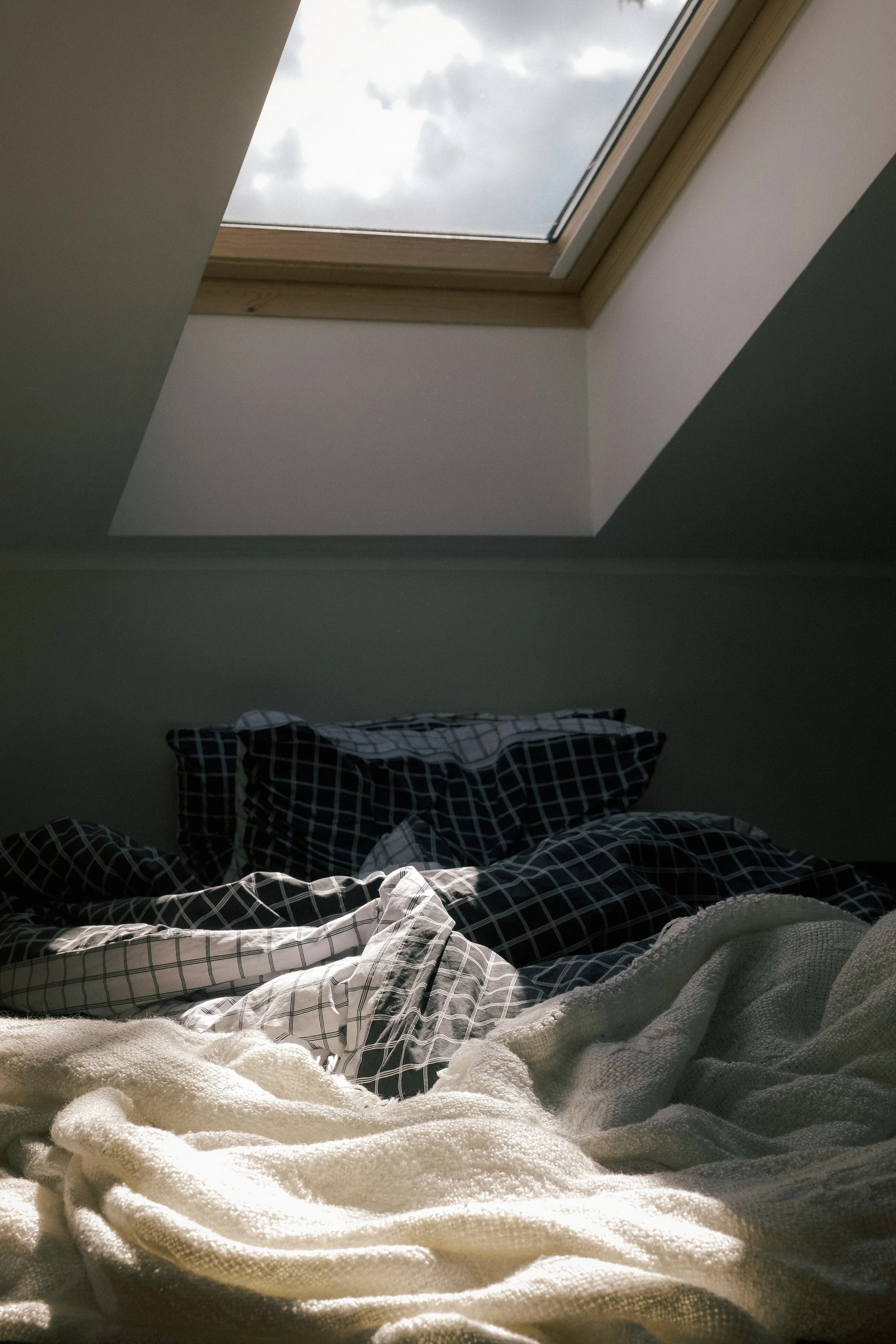 A bedroom with a sloped ceiling and a skylight window showing cloudy sky outside. The bed has a checkered black and white duvet cover and a white fleece blanket in the foreground.