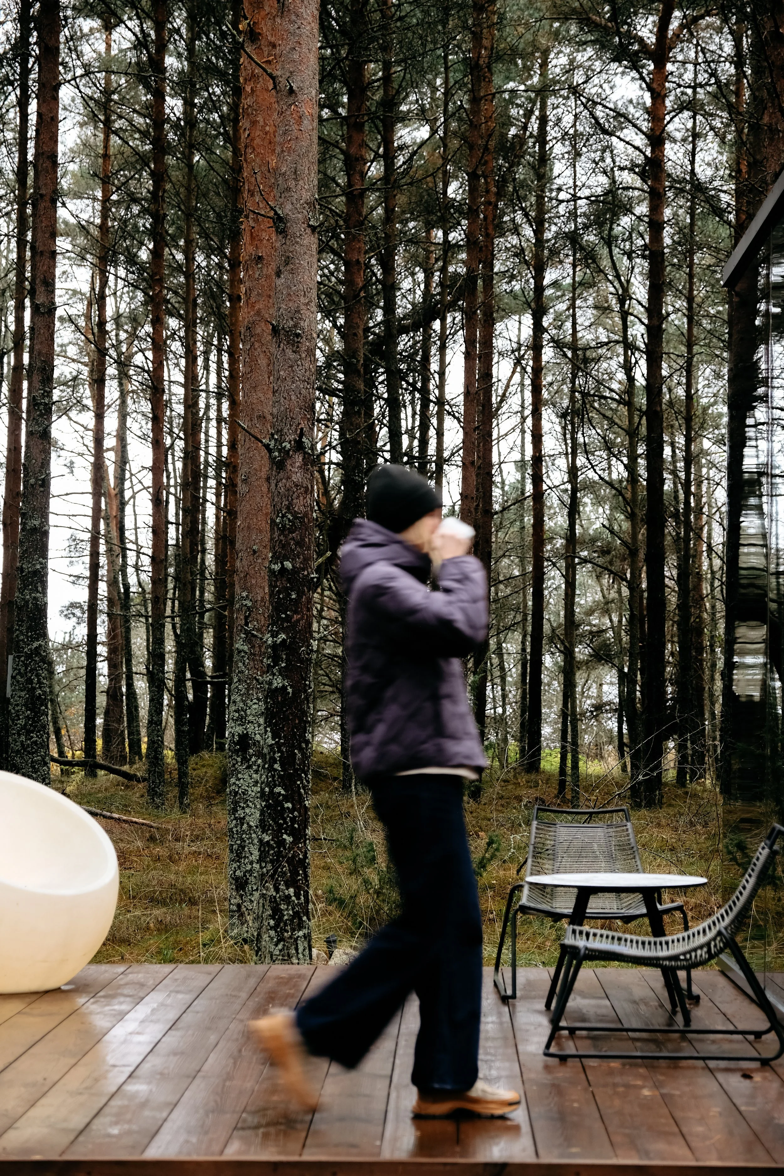 A person walking on a wooden deck in a forested area, wearing a dark jacket, black beanie, and tan boots, with chairs and trees around.