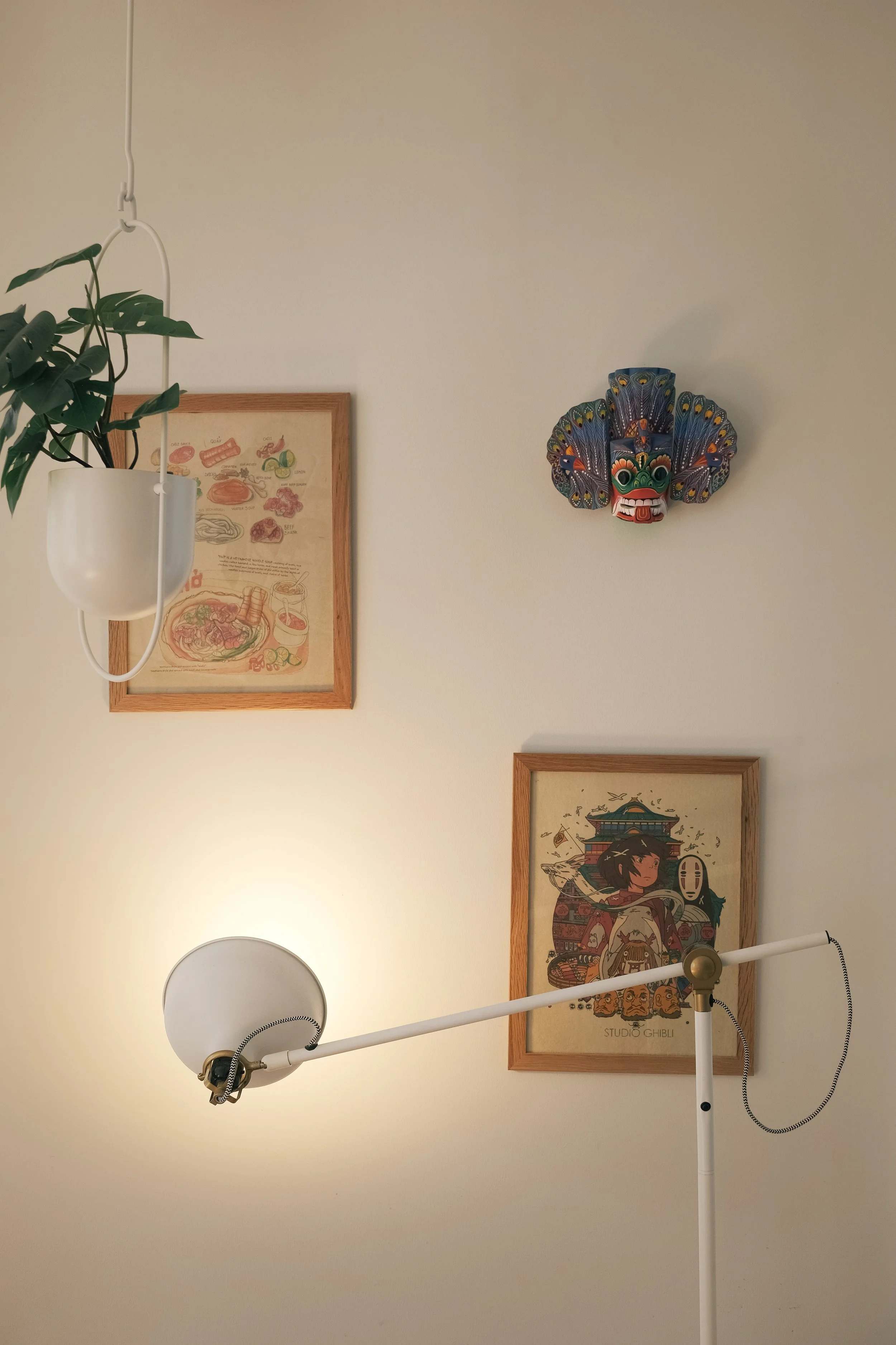 Wall decorated with framed artwork and a colorful mask resembling a bird, illuminated by a white floor lamp and a hanging planter with a green plant.