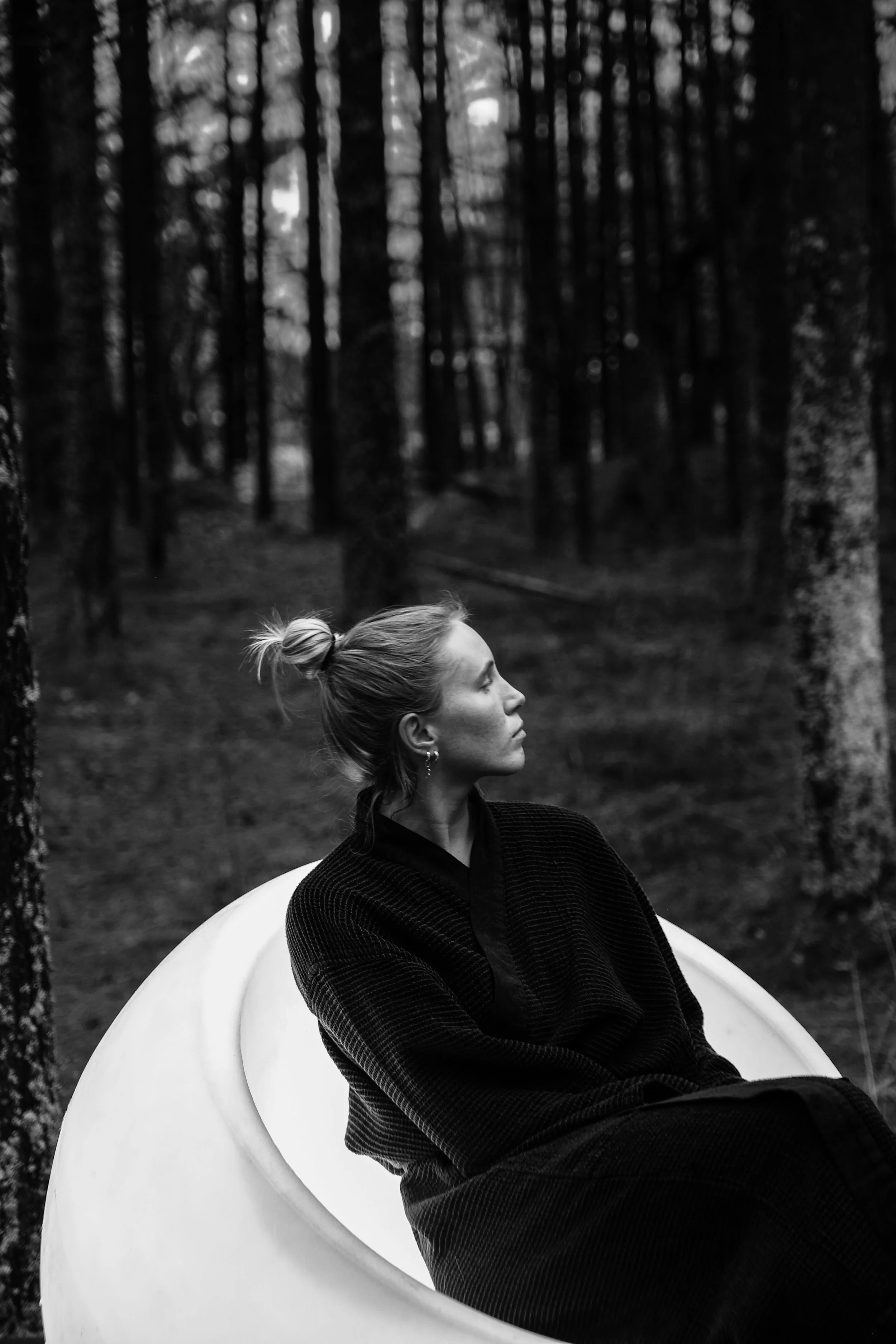 A woman with her hair in a bun, wearing dark clothing, sitting inside a white, curved chair outdoors in a forest with tall trees in the background.