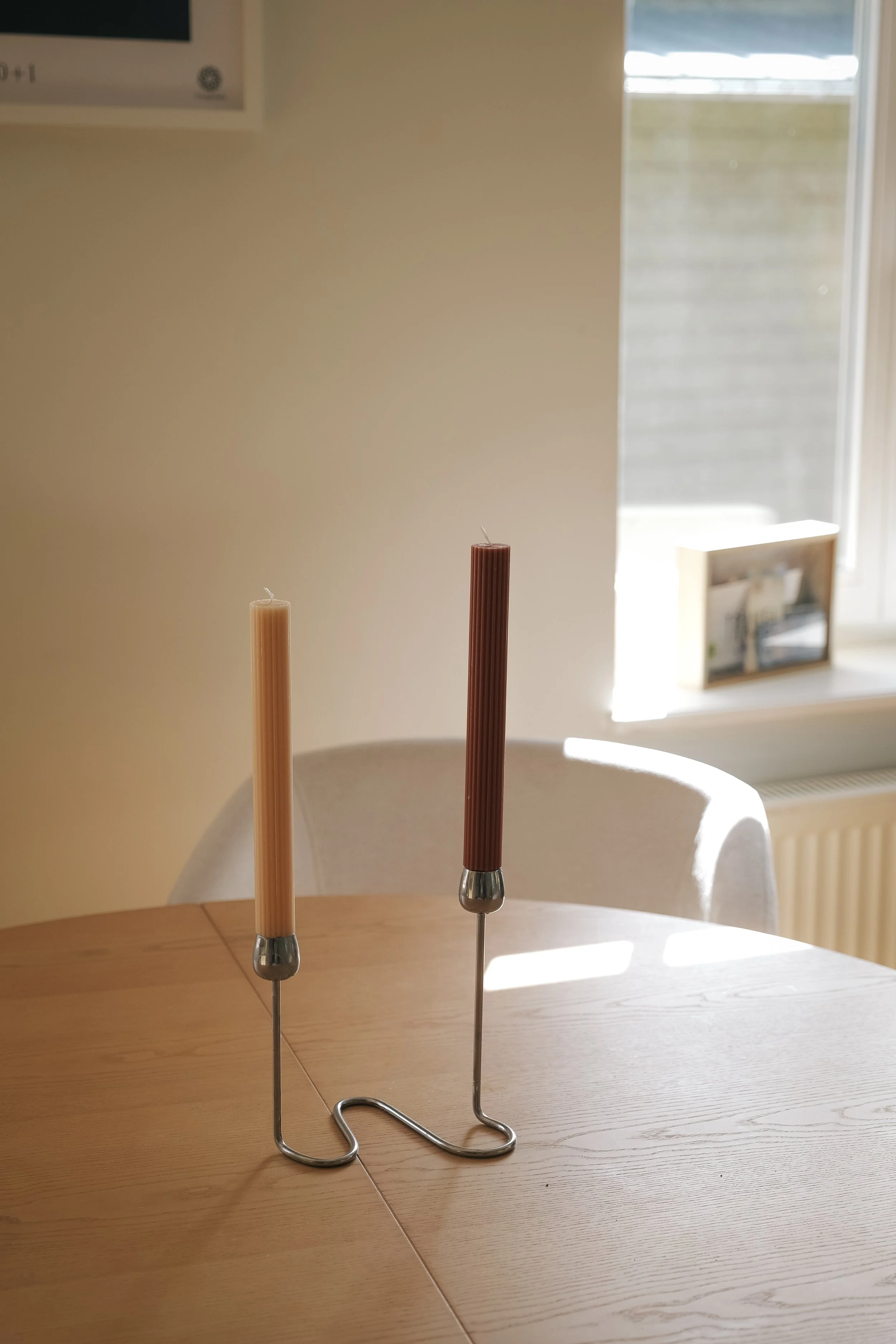 Two candle holders with long, thin candles, one beige and one brown, on a wooden table near a window with sunlight.