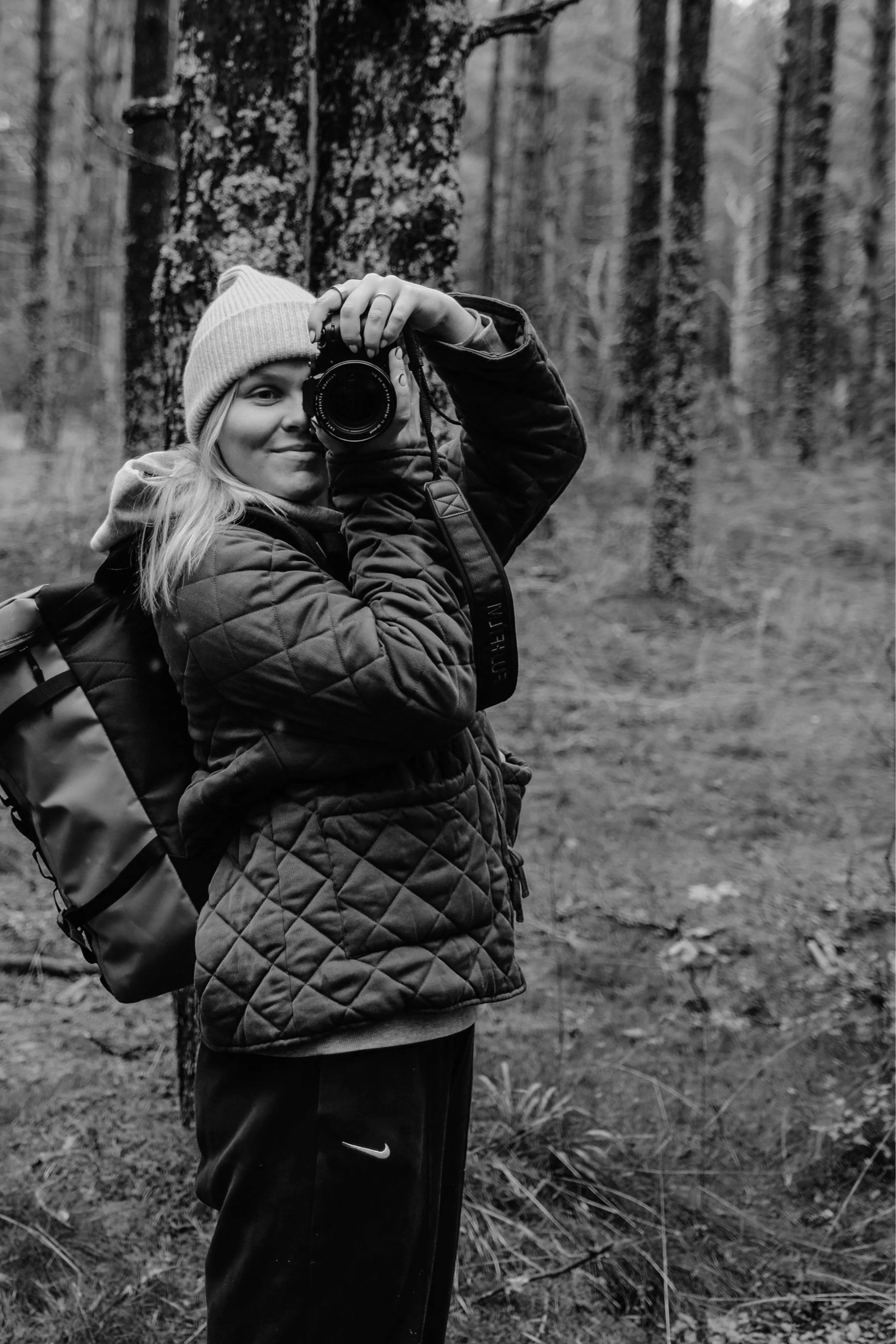 A person wearing a beanie hat and a quilted jacket is holding a camera and taking a selfie in a forest with tall trees and fallen leaves on the ground.