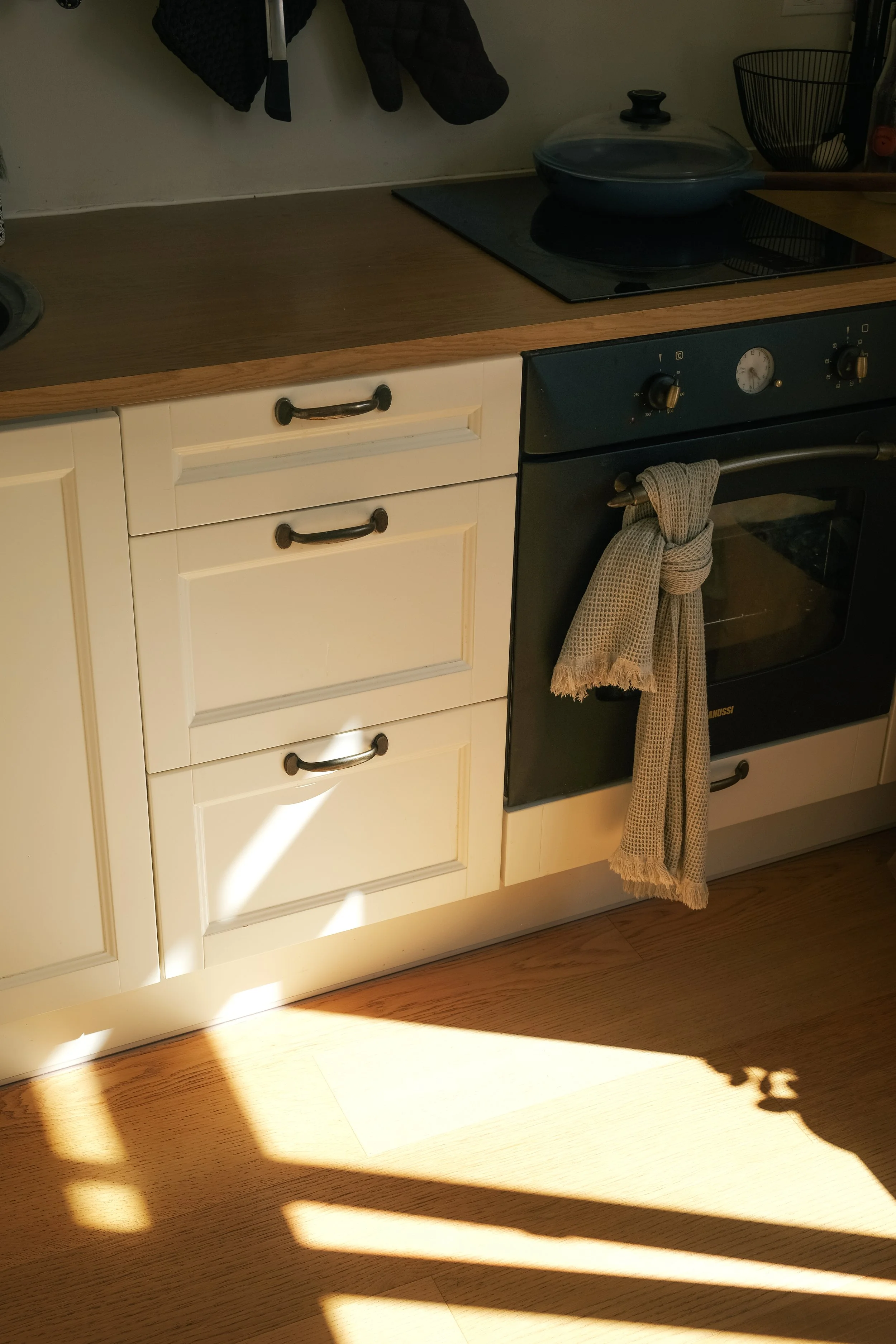 A kitchen countertop with a black stovetop, a glass lid covering a frying pan, and various kitchen utensils. Below the countertop are white cabinets with black handles. A cloth hangs from the oven door handle, and sunlight casts shadows on the wooden floor.