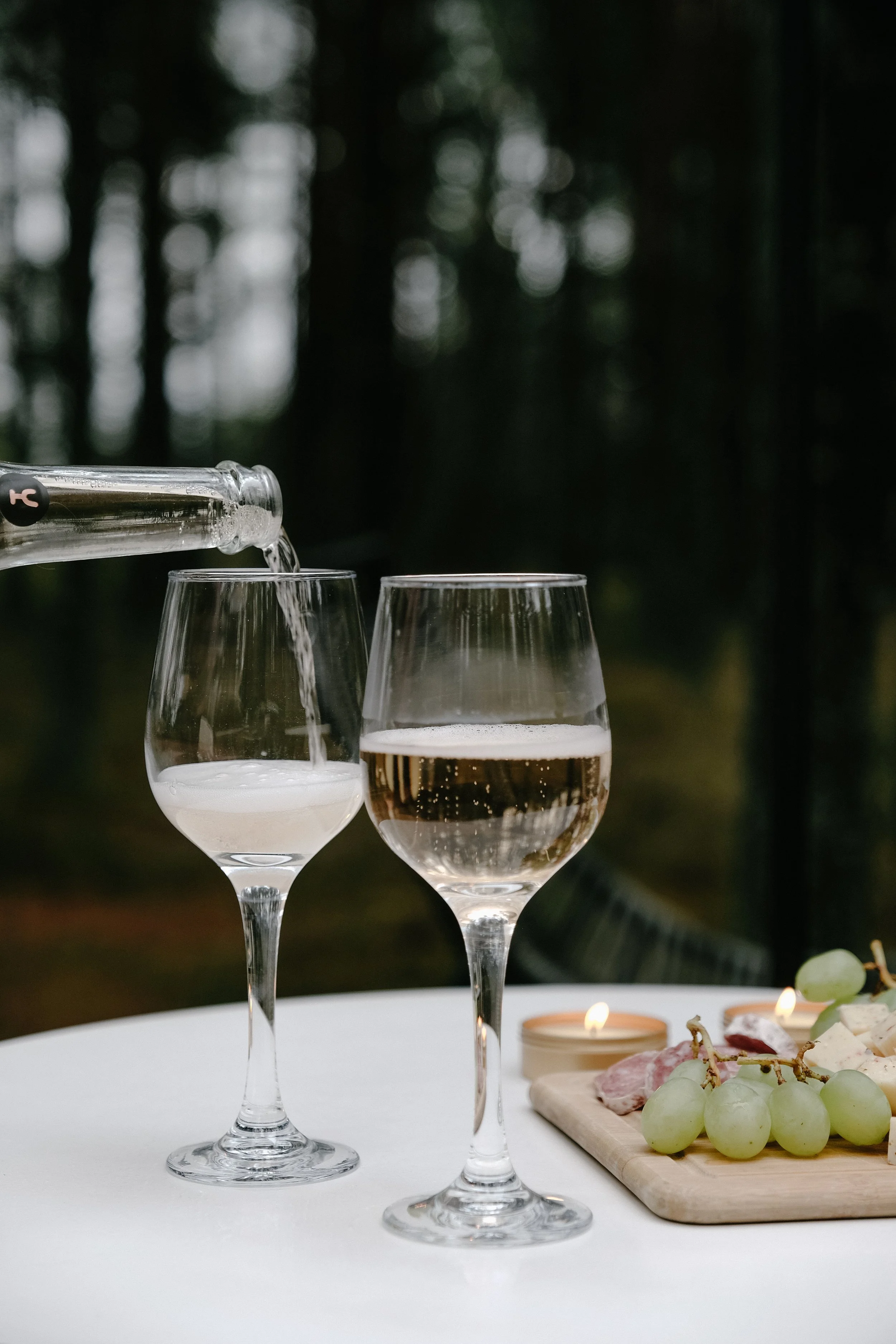 Pouring water from a glass bottle into a wine glass, with two wine glasses in the foreground, on a white table with grapes, cheese, charcuterie, candles, and dessert in a blurred outdoor setting.