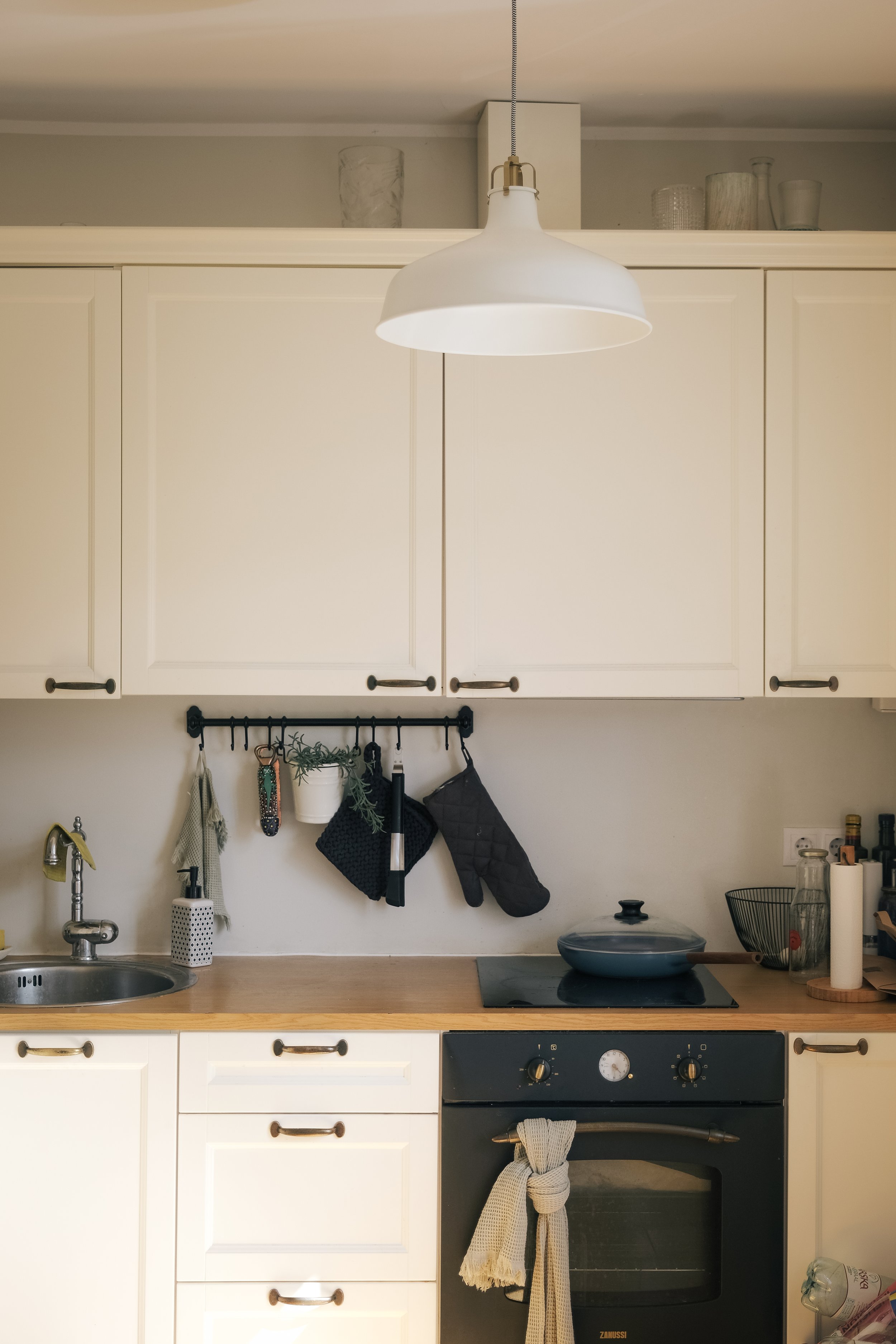 A modern kitchen with white cabinets, a wooden countertop, and a black oven. There's a white pendant light hanging from the ceiling. The backsplash area has a black metal rail with hanging kitchen items including a potted plant, dish towels, and oven mitts. The kitchen also features a sink with a gold faucet and various kitchen utensils.