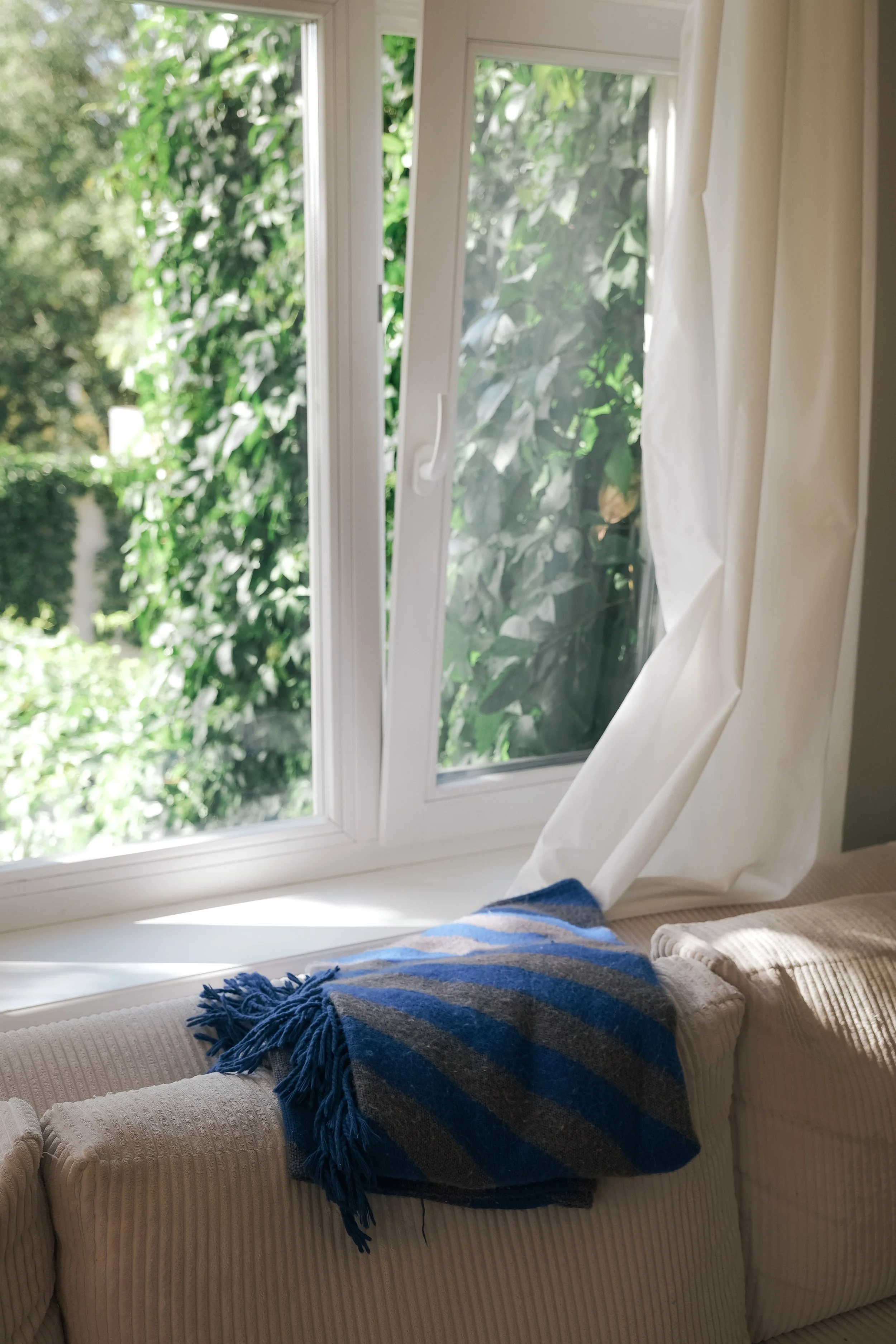 View of a window with white curtains and greenery outside. A beige textured sofa has a blue and gray striped blanket and a beige cushion on it.