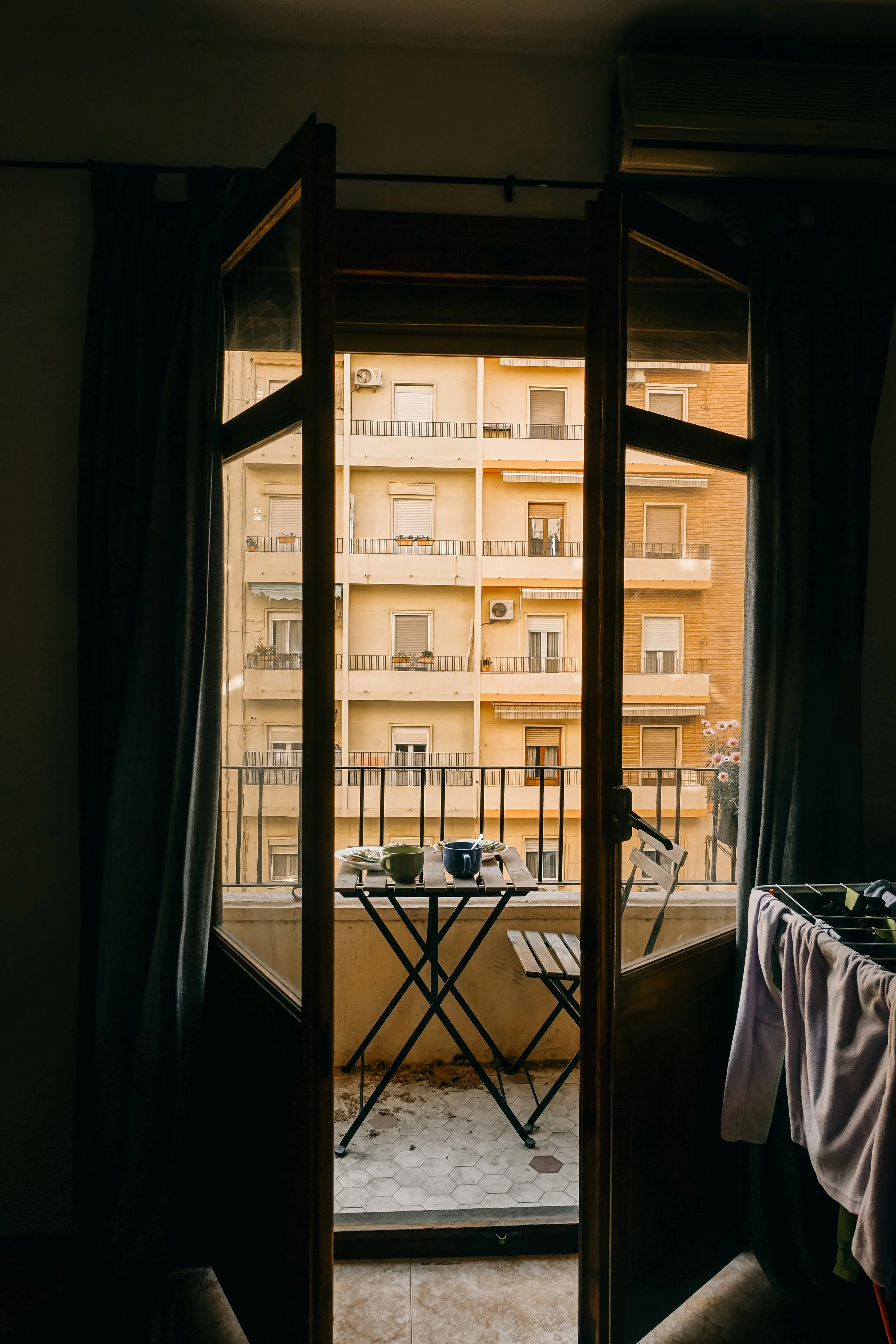 View of a balcony from inside an apartment, with two cups on a small table, a folding chair, and laundry hanging on a clothesline; building with balconies and windows in the background.
