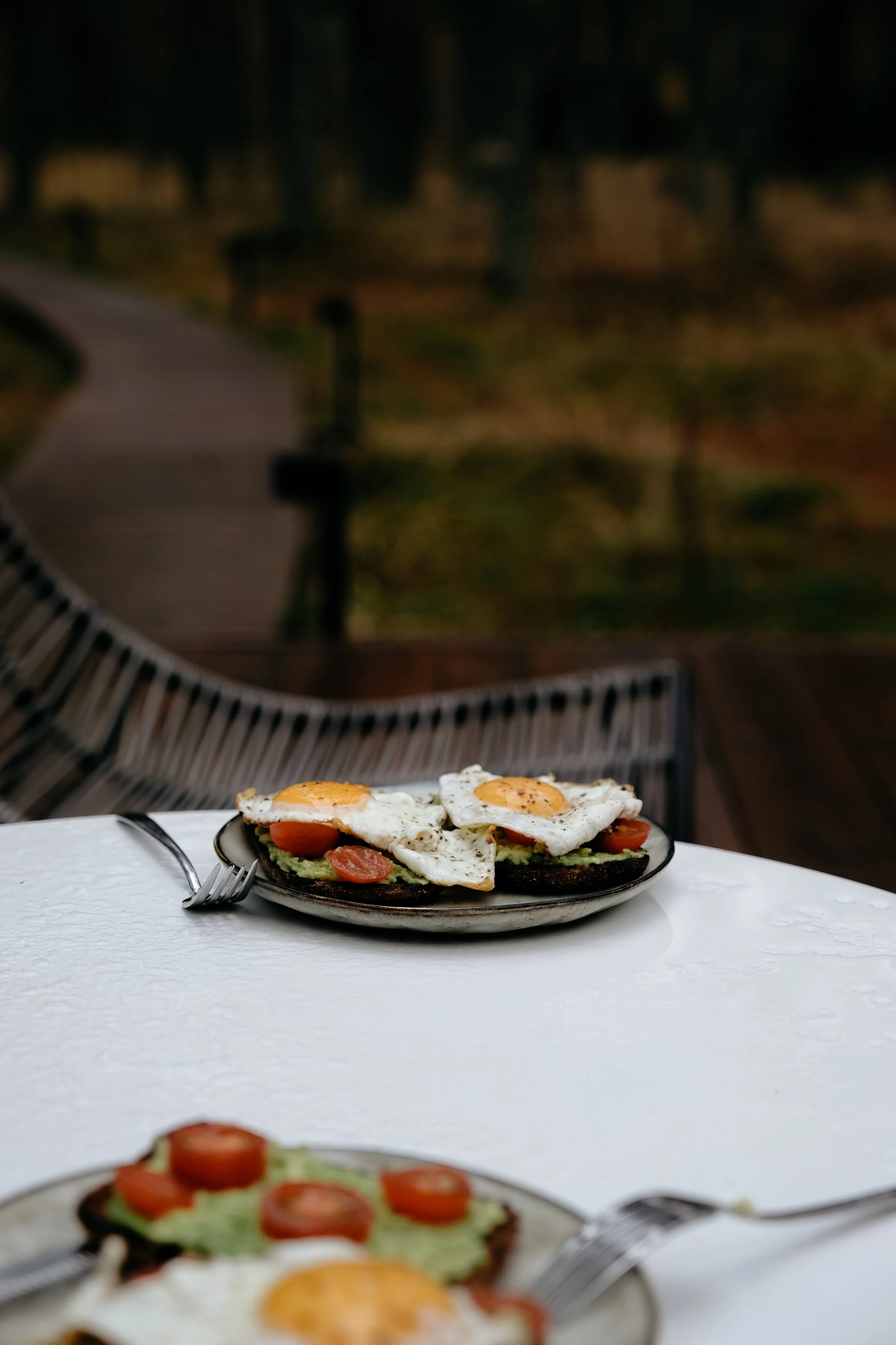 Plate of avocado toast topped with cherry tomatoes, and two fried eggs with sunny-side up, on a white tablecloth. In the background, an outdoor pathway with trees and a bench.