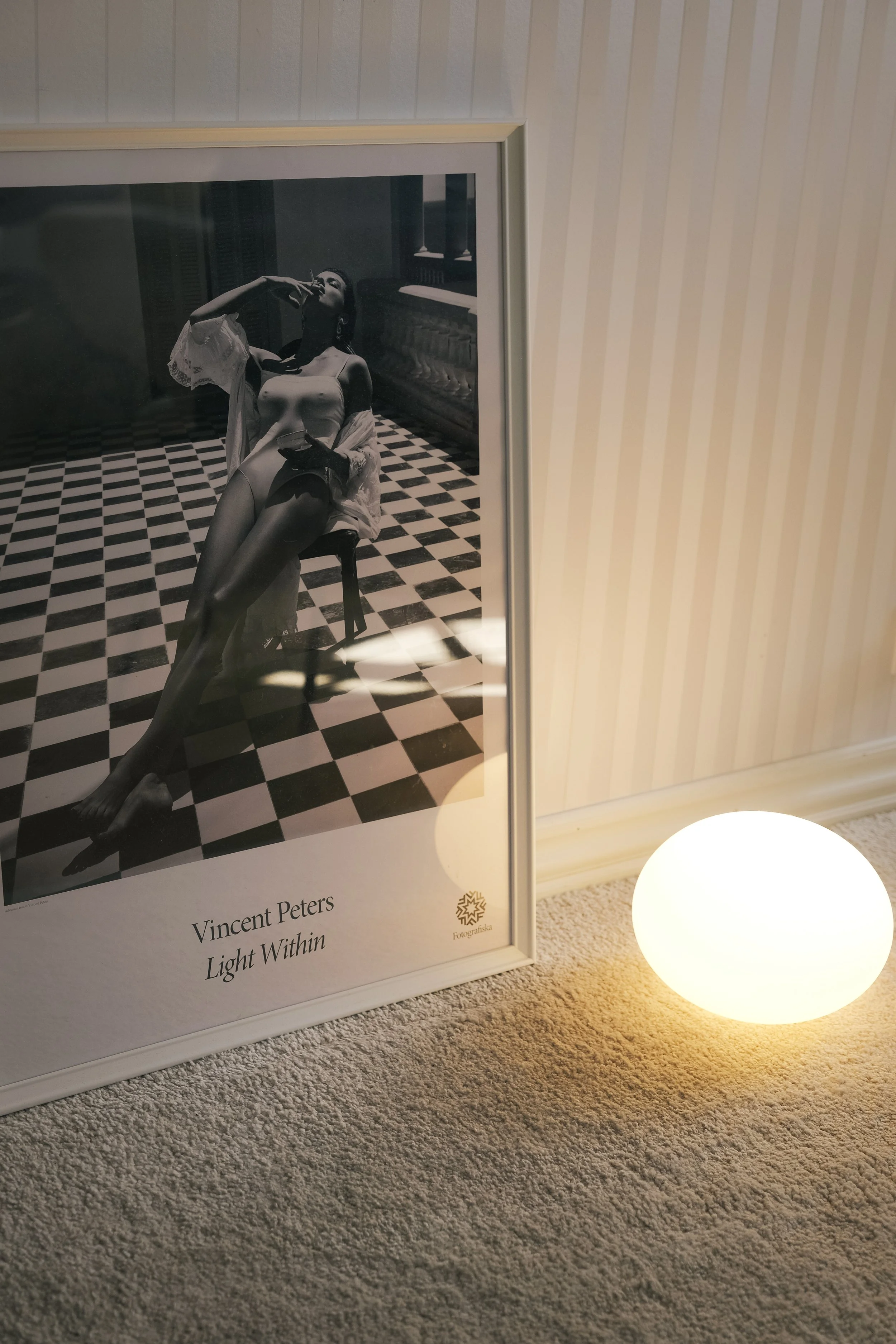 A framed black and white photograph of a woman sitting on a chair in a room with checkered floor, labeled with text: Vincent Peters Light Within. A glowing spherical light is on the floor in front of the frame.