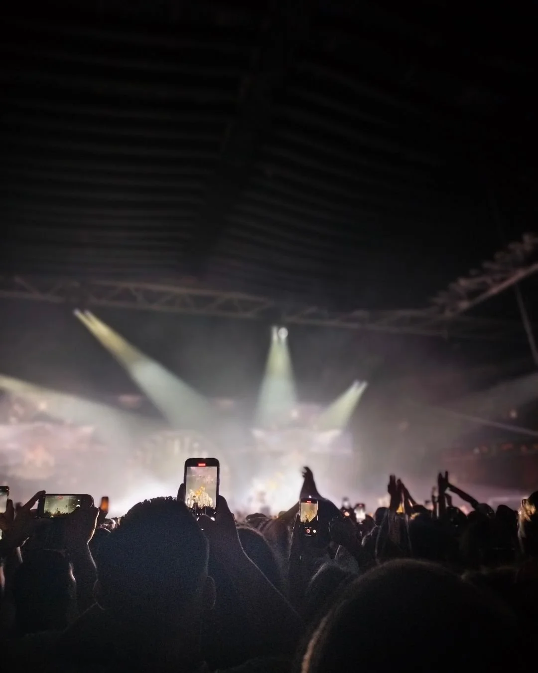 Crowd at a concert with people taking photos or videos with their phones, stage with lights in the background, inside a large indoor venue.
