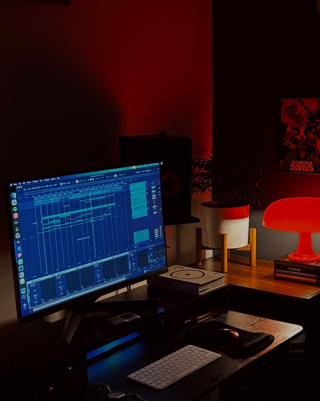A music production workspace with a computer monitor displaying digital audio workstation software, a keyboard, a mouse, and sound equipment, with a warm red and orange ambient lighting.