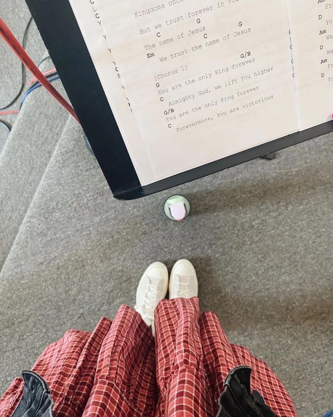 A person wearing red checkered skirt and white sneakers standing on gray carpet in front of a music stand holding sheet music with song lyrics and chords, next to an electronic device with cables.