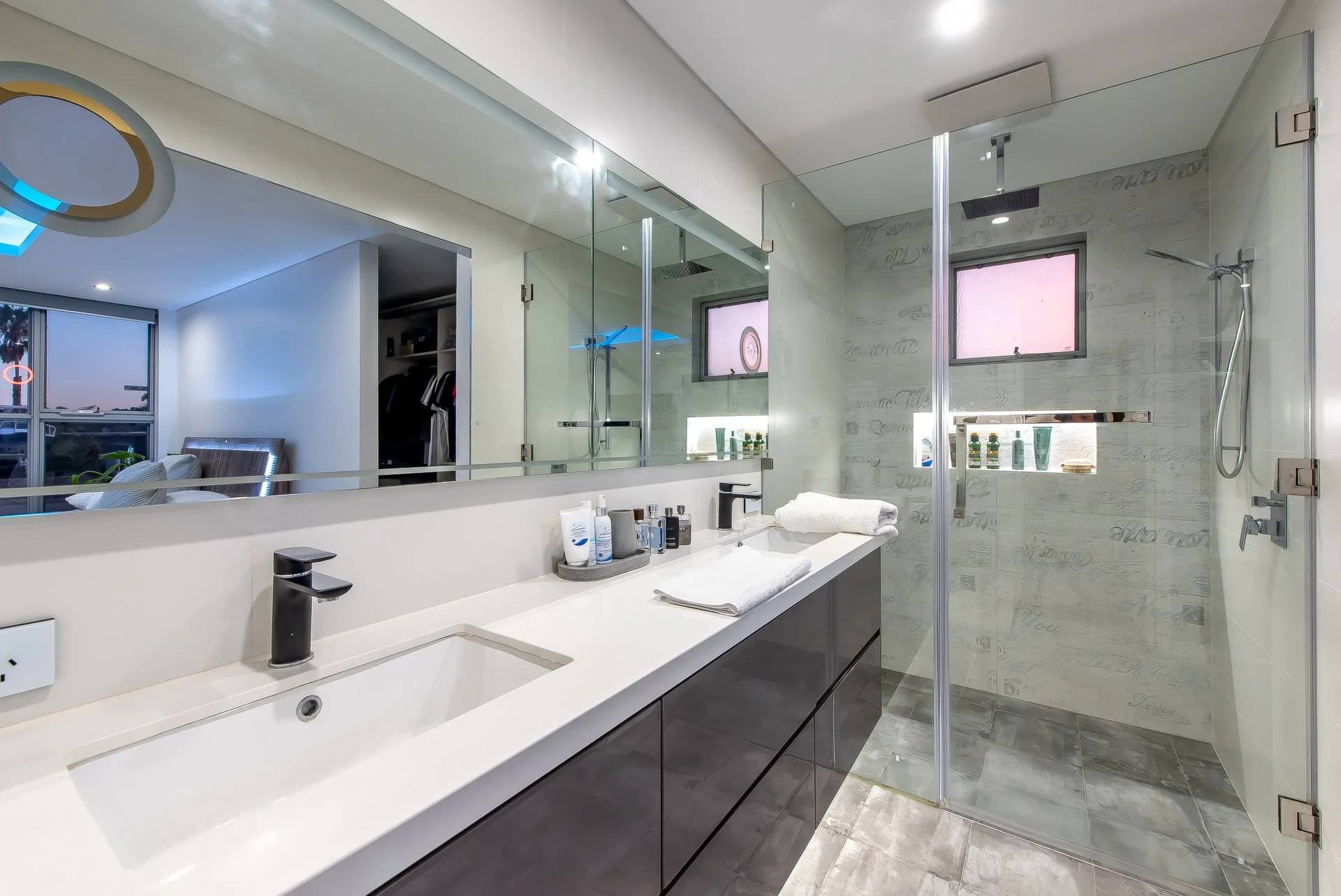 Modern bathroom with large mirror, white vanity, black faucet, towels, shower with glass door, gray tiles, small window, toiletries on shelf, closet with clothes, and living room with bed and window in background.