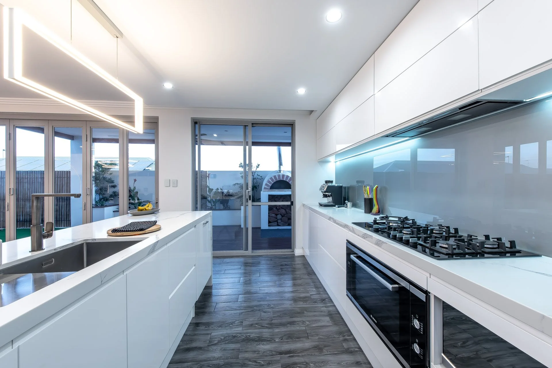 Modern kitchen with white cabinets, a stove, and a kitchen island with a sink. Sliding glass doors lead to an outdoor area with a pizza oven and patio furniture.