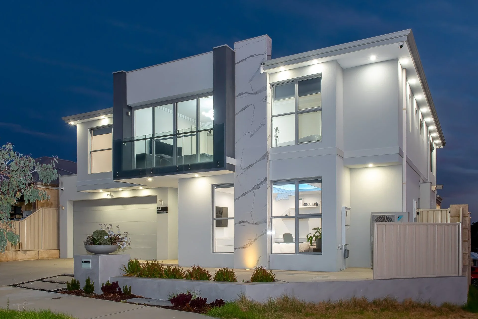 Modern white multi-story house with large windows and a balcony, lit up at night, surrounded by a landscaped front yard with plants and a fence.