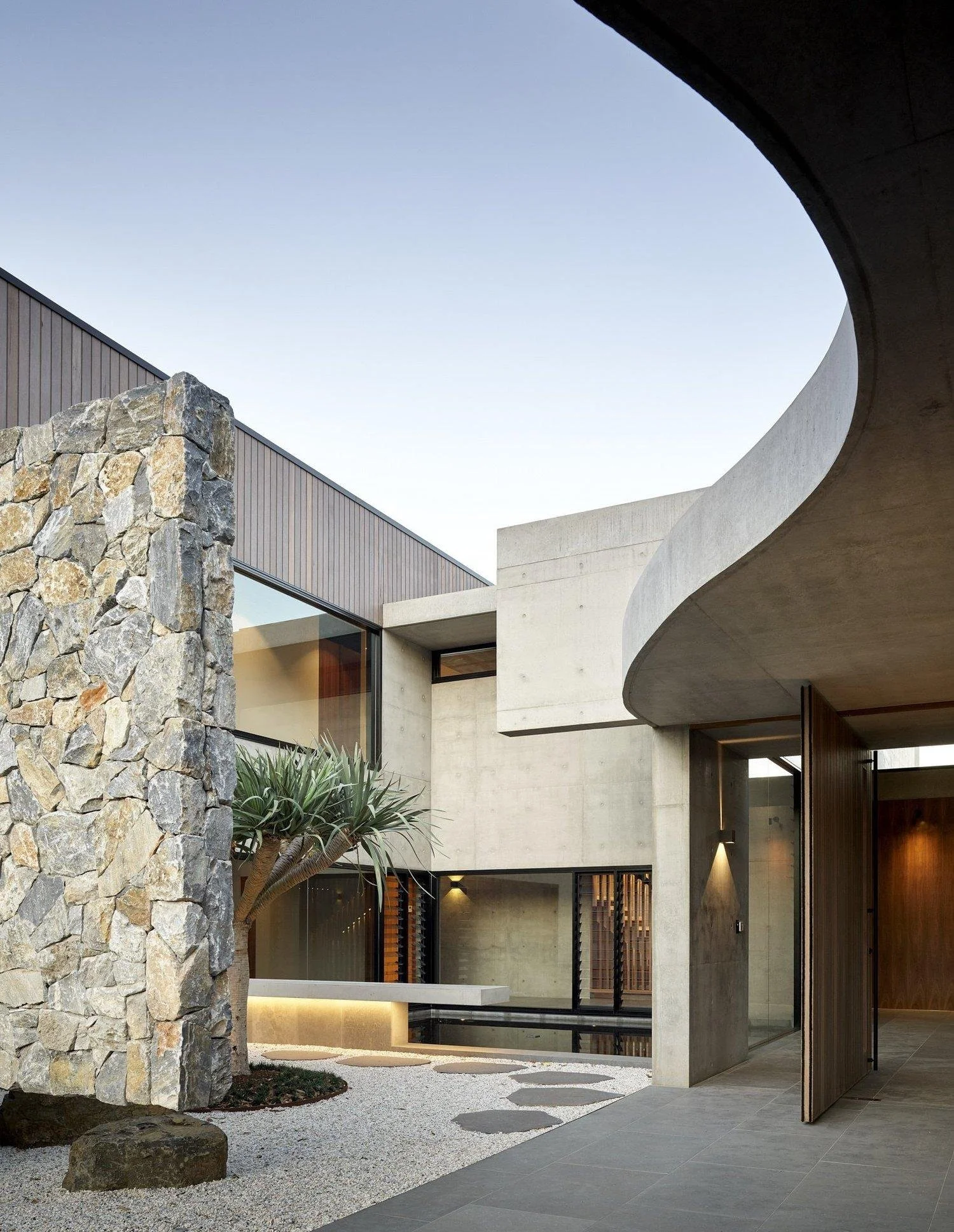 Modern house exterior with stone wall, large windows, and curved concrete roof, surrounded by a gravel pathway and desert plants under a clear sky.