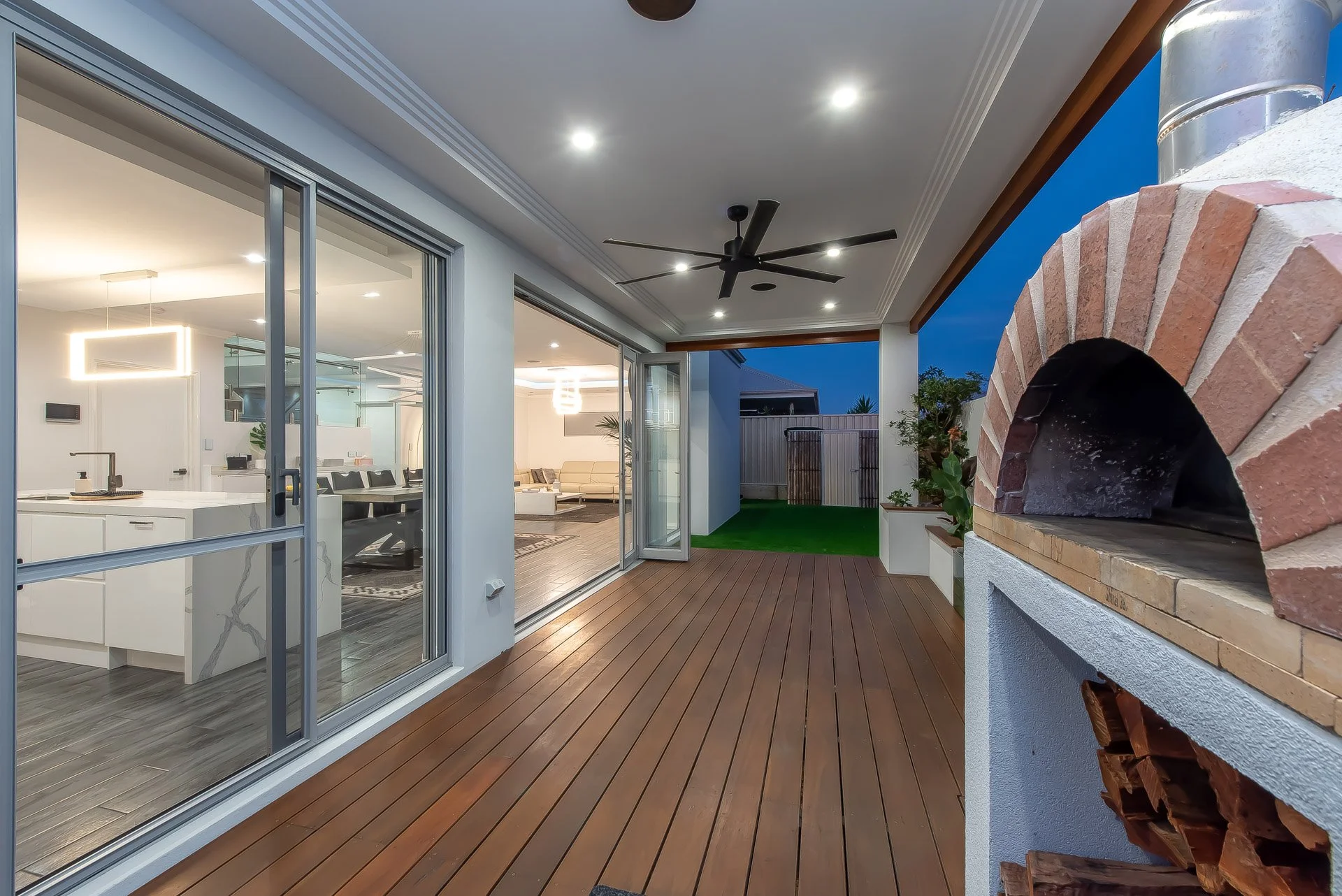 Covered outdoor patio with wood flooring, a ceiling fan, and a built-in brick and concrete pizza oven on the right. Large glass sliding doors lead to an interior living space with modern furnishings. The sky is dark, indicating evening.