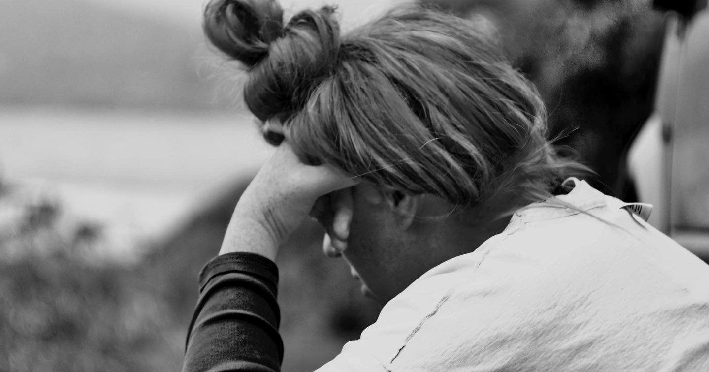 A woman sitting outdoors with her head resting on her hand, looking downward, appearing upset or contemplative.