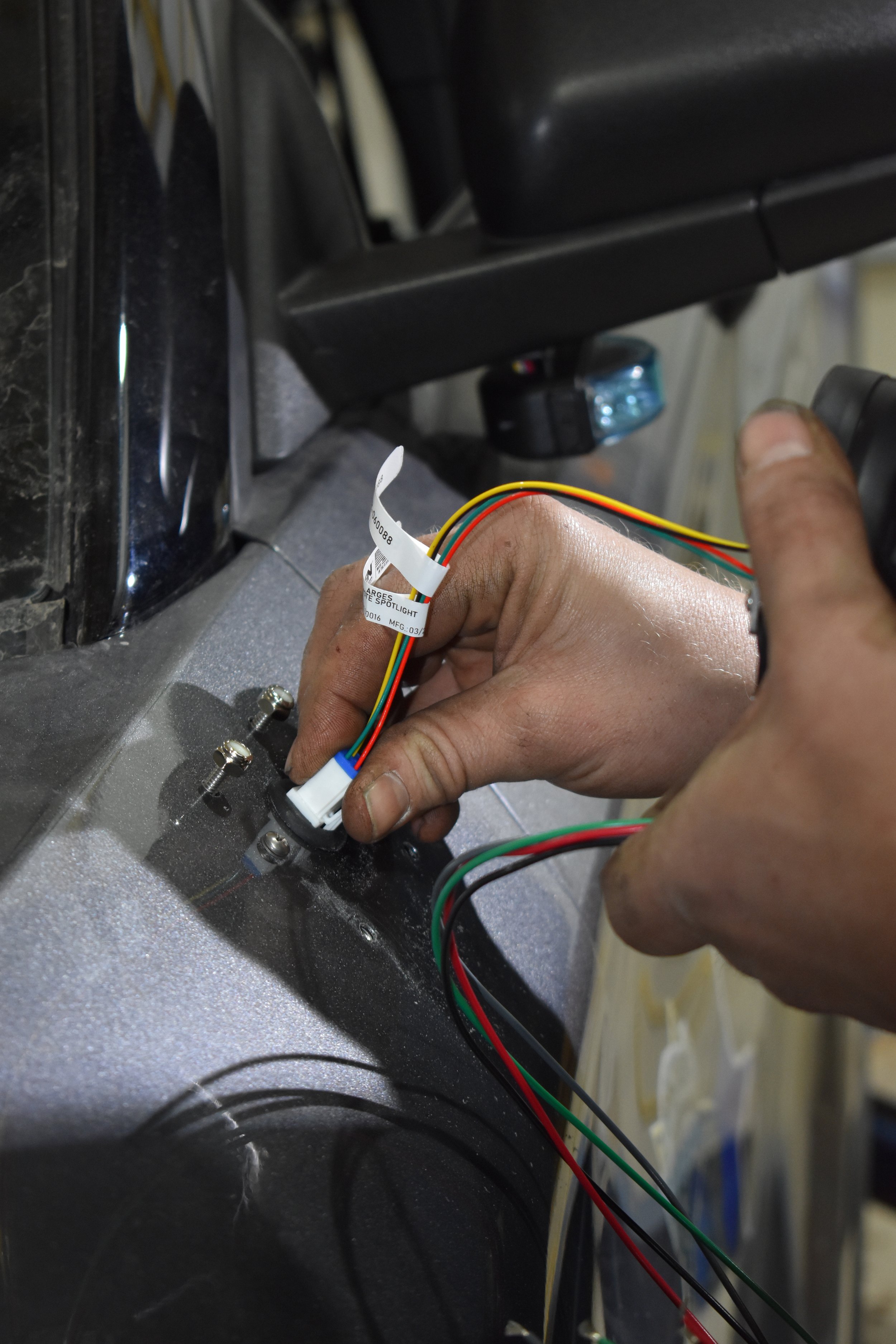 A person holding an electronic wiring connector with multiple colorful wires, connected to a metal surface with screws, in a repair or installation setting.